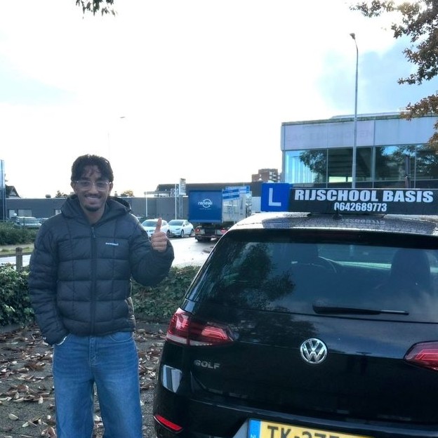 Jonge man in grijze jas geeft duimen omhoog naast een zwarte Volkswagen Golf met een rijschoolbord en een geel Nederlands kenteken.