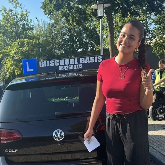 Vrouw lacht en toont duim omhoog naast zwarte Volkswagen Golf met 'Rijschool Basis' bord op het dak.