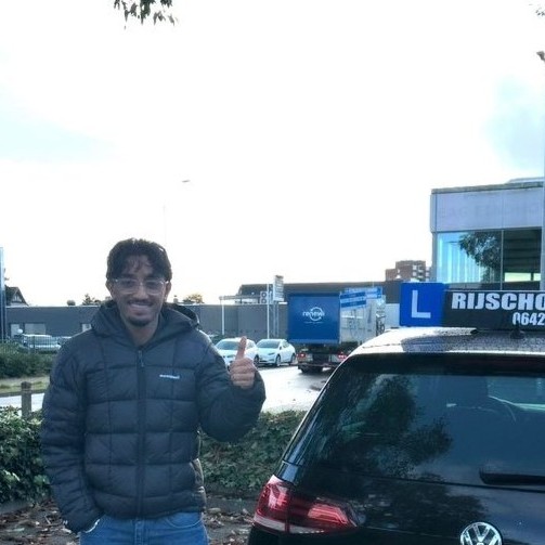 Jonge man met bril en donkere jas steekt duim op naast een zwarte auto met rijschoolbord op het dak.