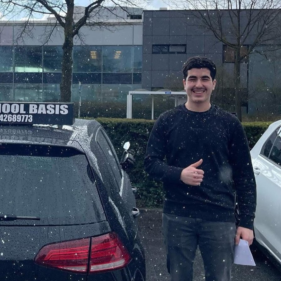 Jonge man met zwarte trui staat lachend naast een zwarte auto met een 'School Basis'-bord buiten op een bewolkte dag.