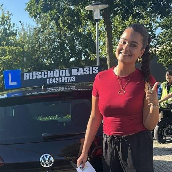 Jonge vrouw die glimlacht en een duim omhoog steekt naast een zwarte Volkswagen met een bord 'Rijschool Basis'.