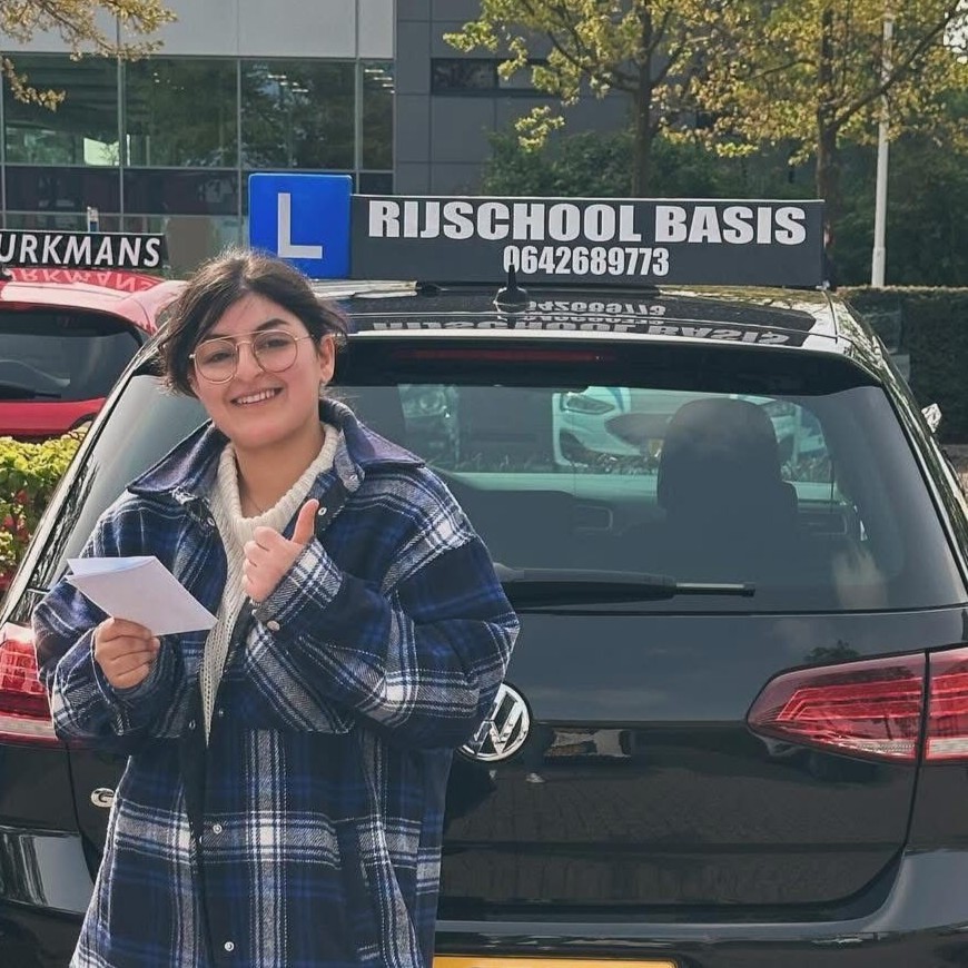 Vrouw met bril en geruite jas glimlacht en toont duim omhoog voor een zwarte auto met bord 'Rijschool Basis'.