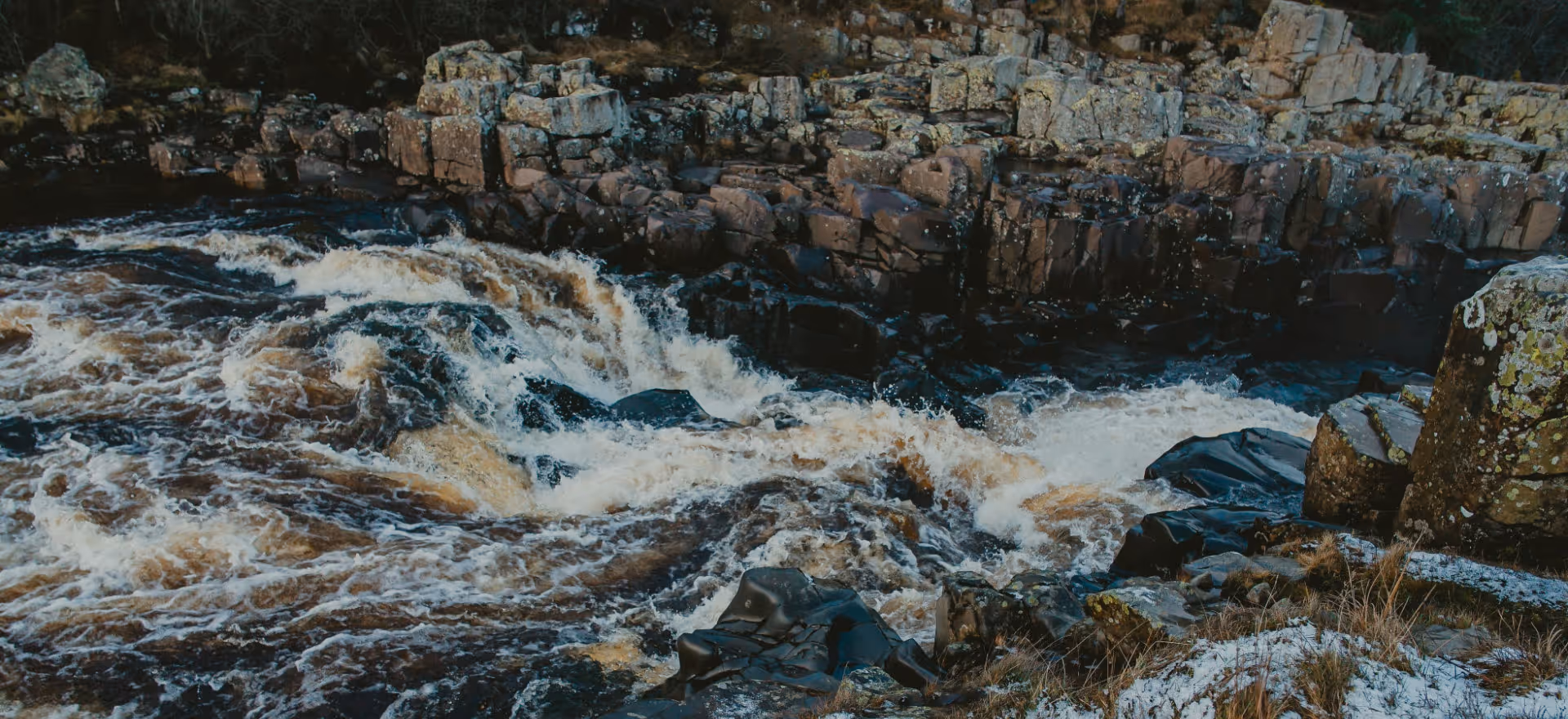 Fast-flowing river cascade surrounded by rugged rocks with a patch of snow-covered ground.