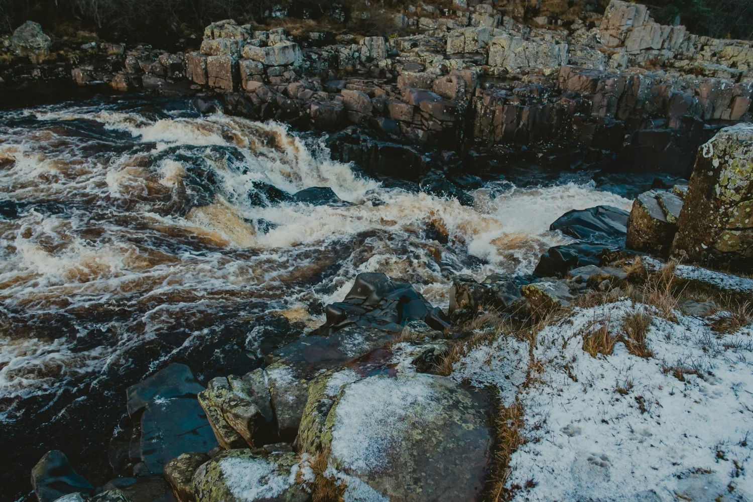 Fast-flowing river cascade surrounded by rugged rocks with a patch of snow-covered ground.