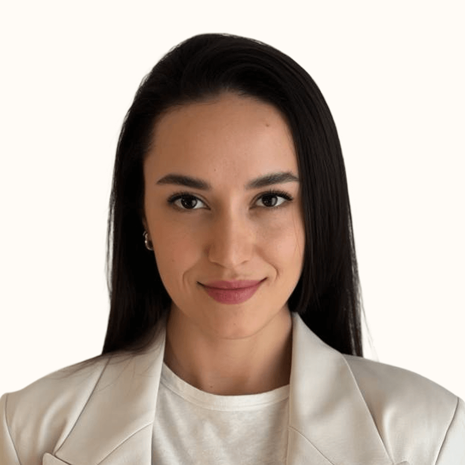 Confident young woman with long dark hair wearing a white blazer and smiling slightly against a light background.