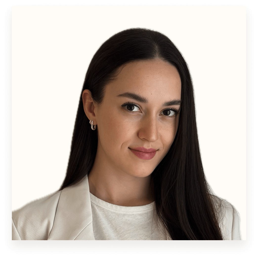 Portrait of a woman with long dark hair, wearing a white blazer and white top, smiling slightly against a plain light background.
