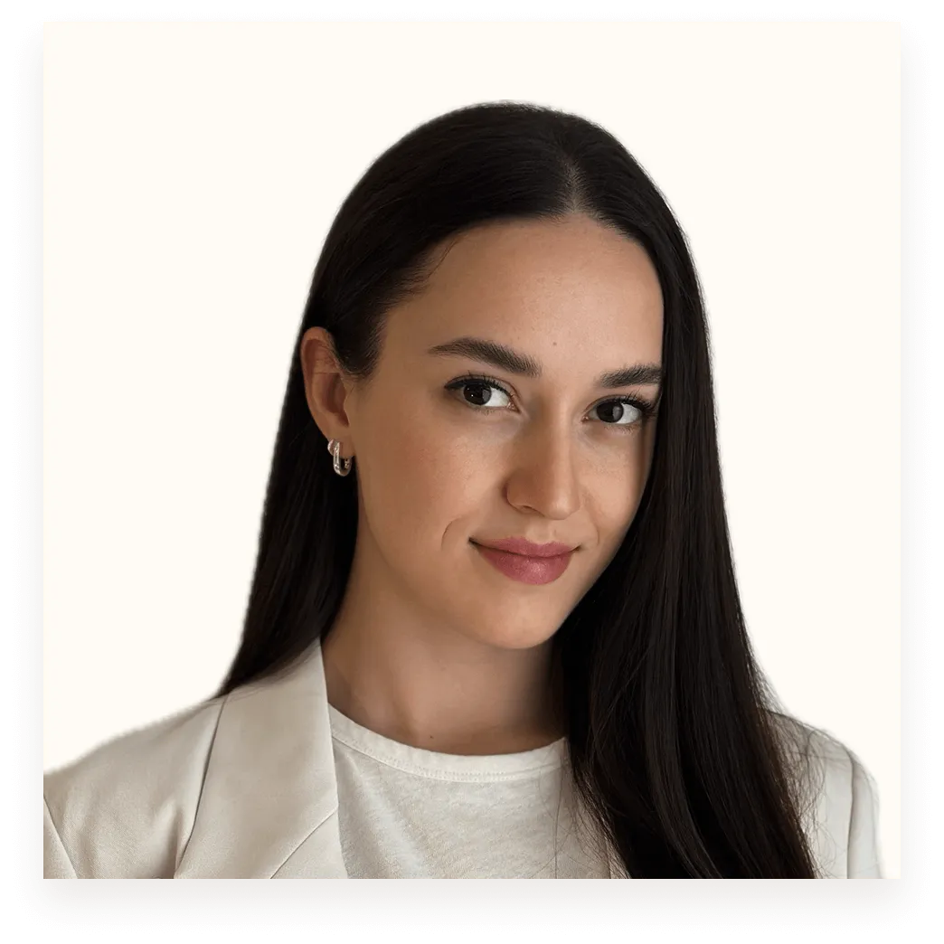 Portrait of a woman with long dark hair, wearing a white blazer and white top, smiling slightly against a plain light background.