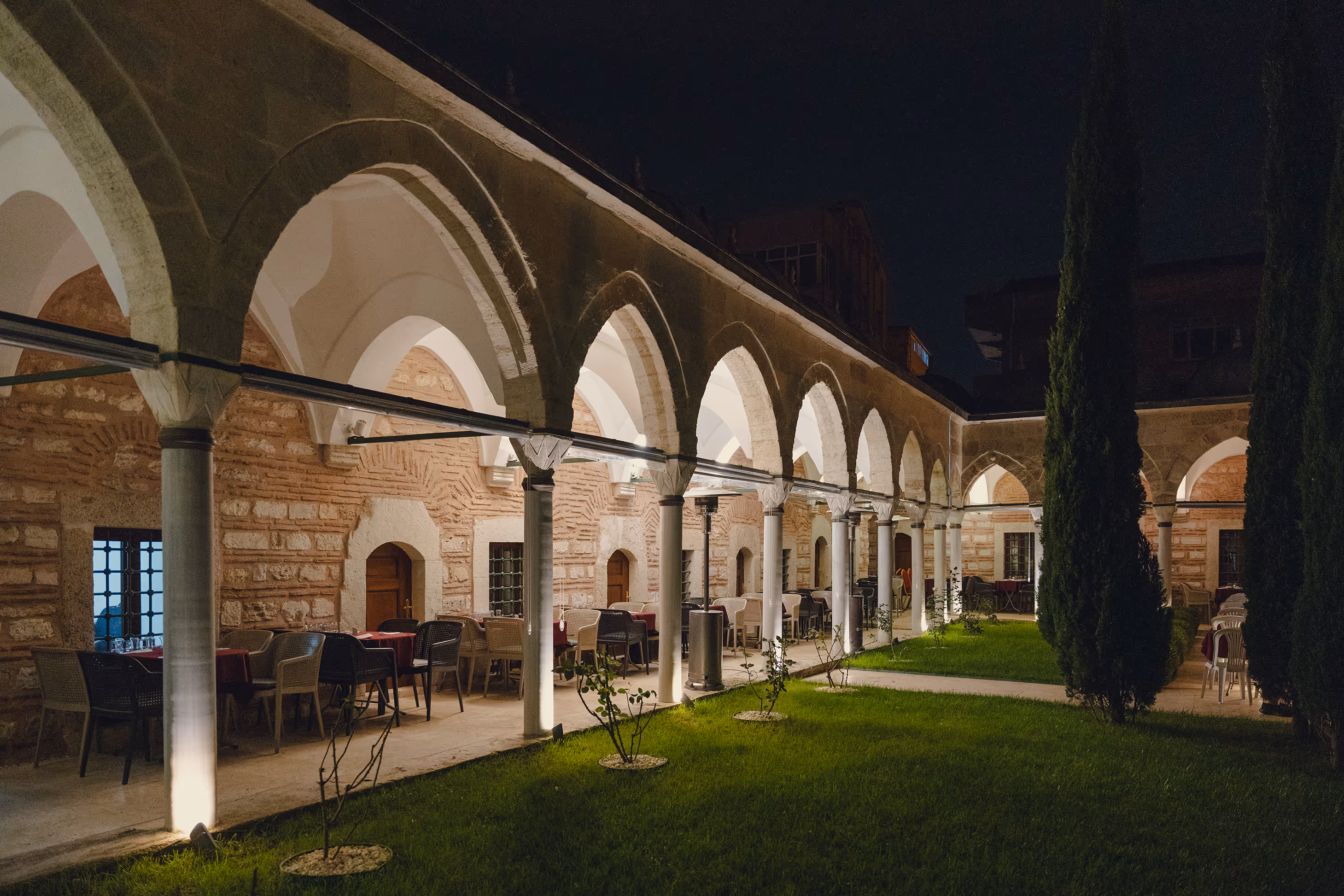 Night view of a courtyard with stone arches, outdoor dining tables, and green lawn with small trees.