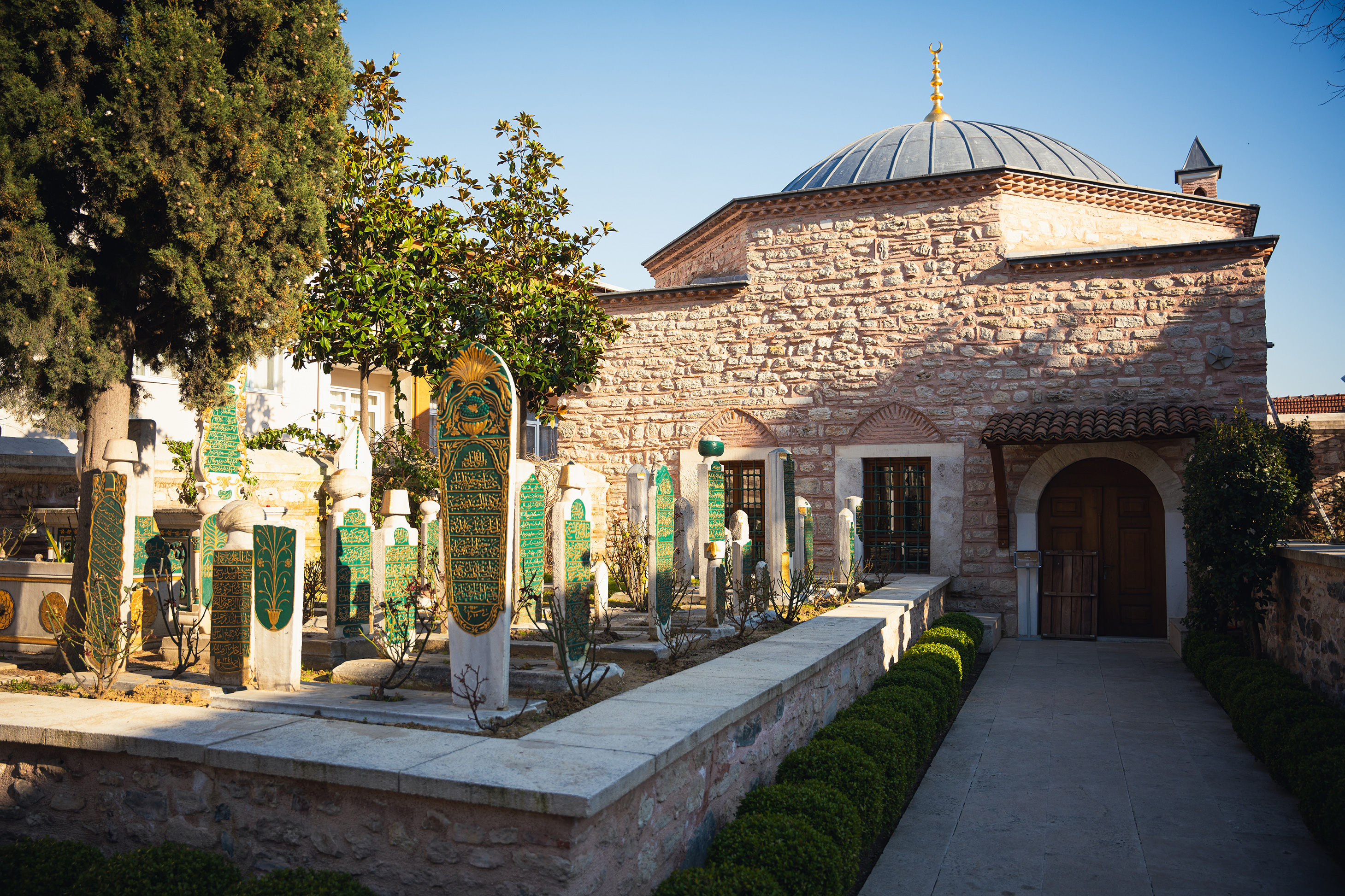 Historic stone mausoleum with a domed roof and an adjacent graveyard featuring ornate green and gold headstones.