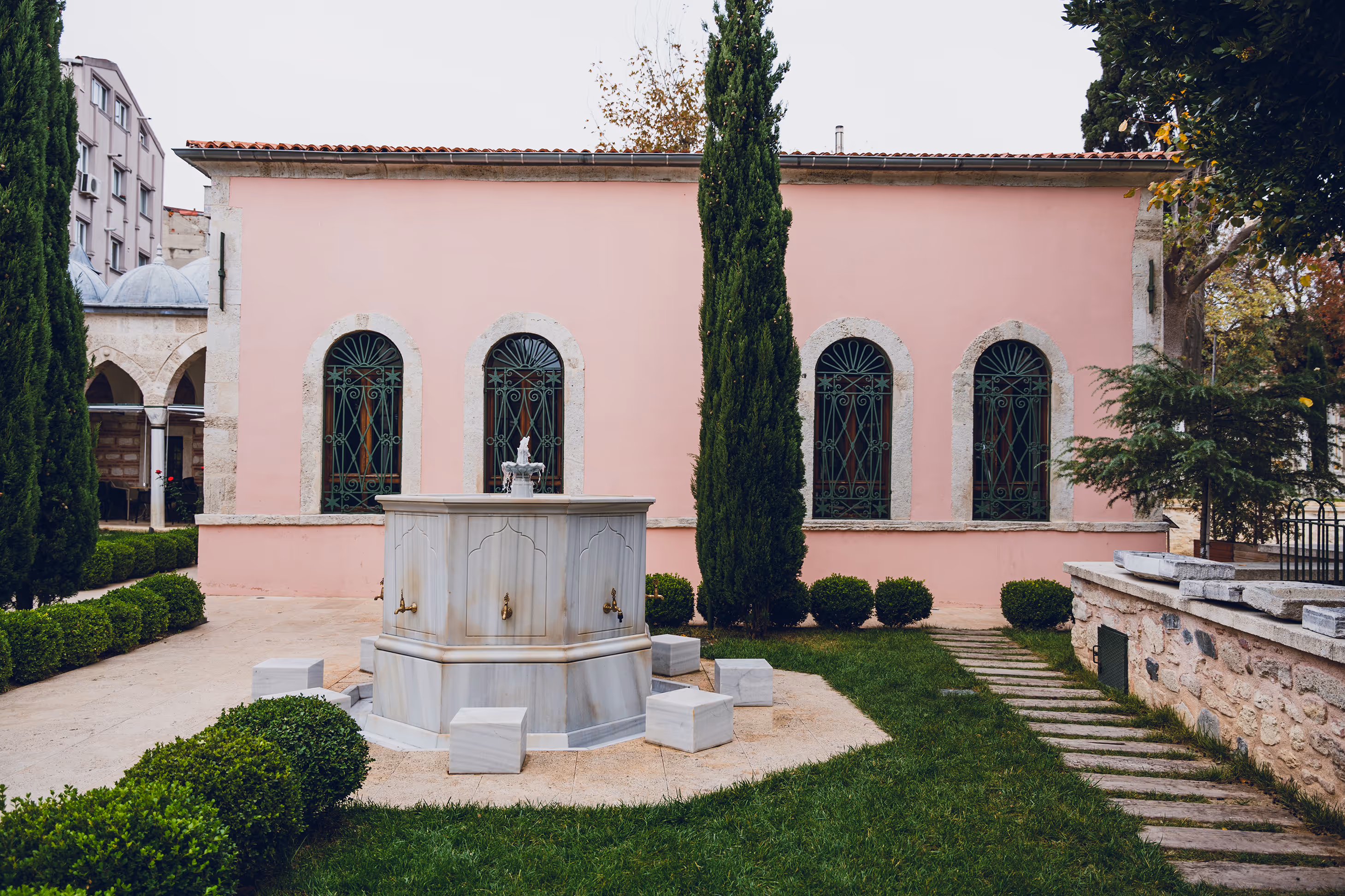 Marble fountain with brass taps and small stone seats in a garden courtyard in front of a pink building with arched windows and green iron grills.