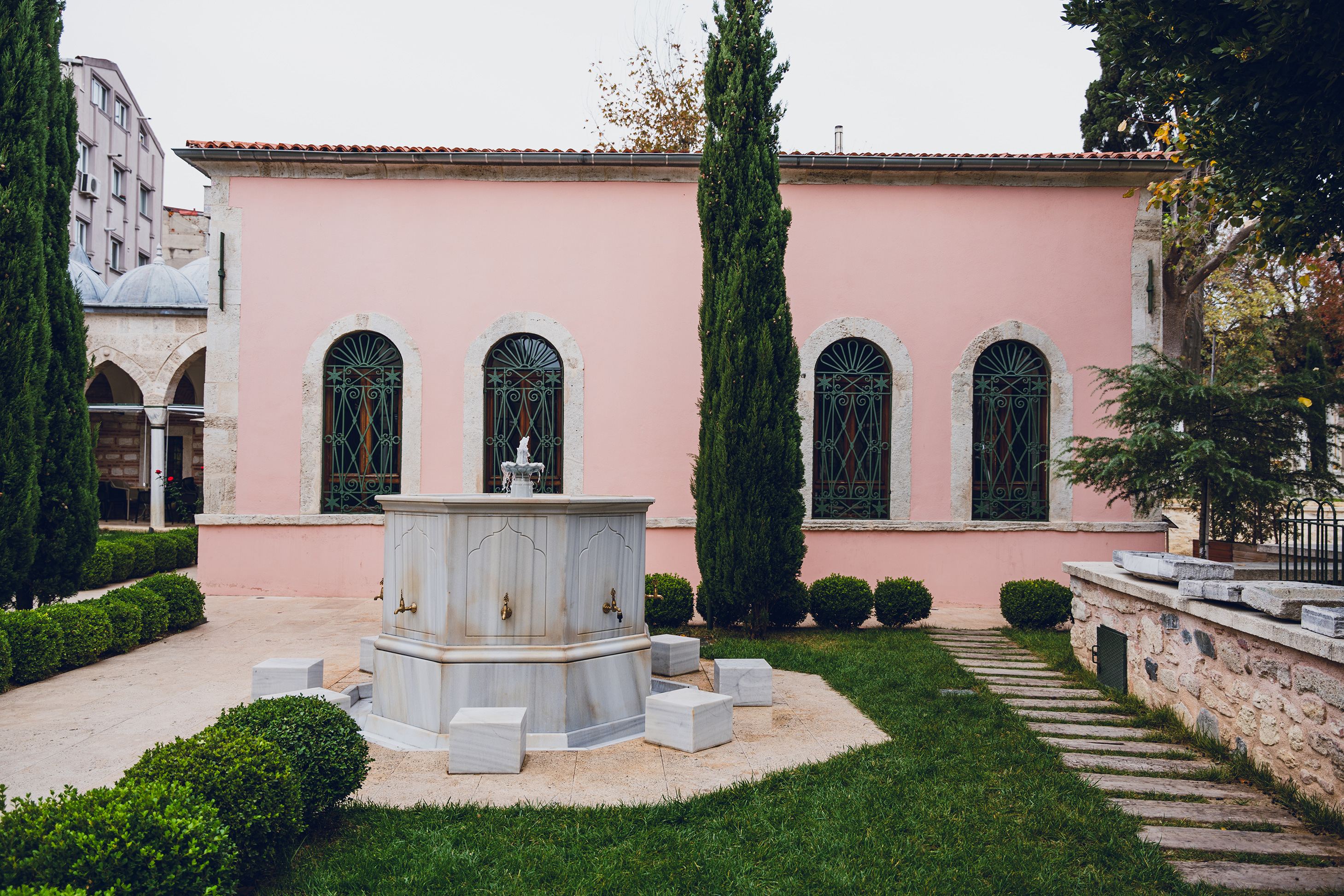 Marble fountain with brass taps and small stone seats in a garden courtyard in front of a pink building with arched windows and green iron grills.