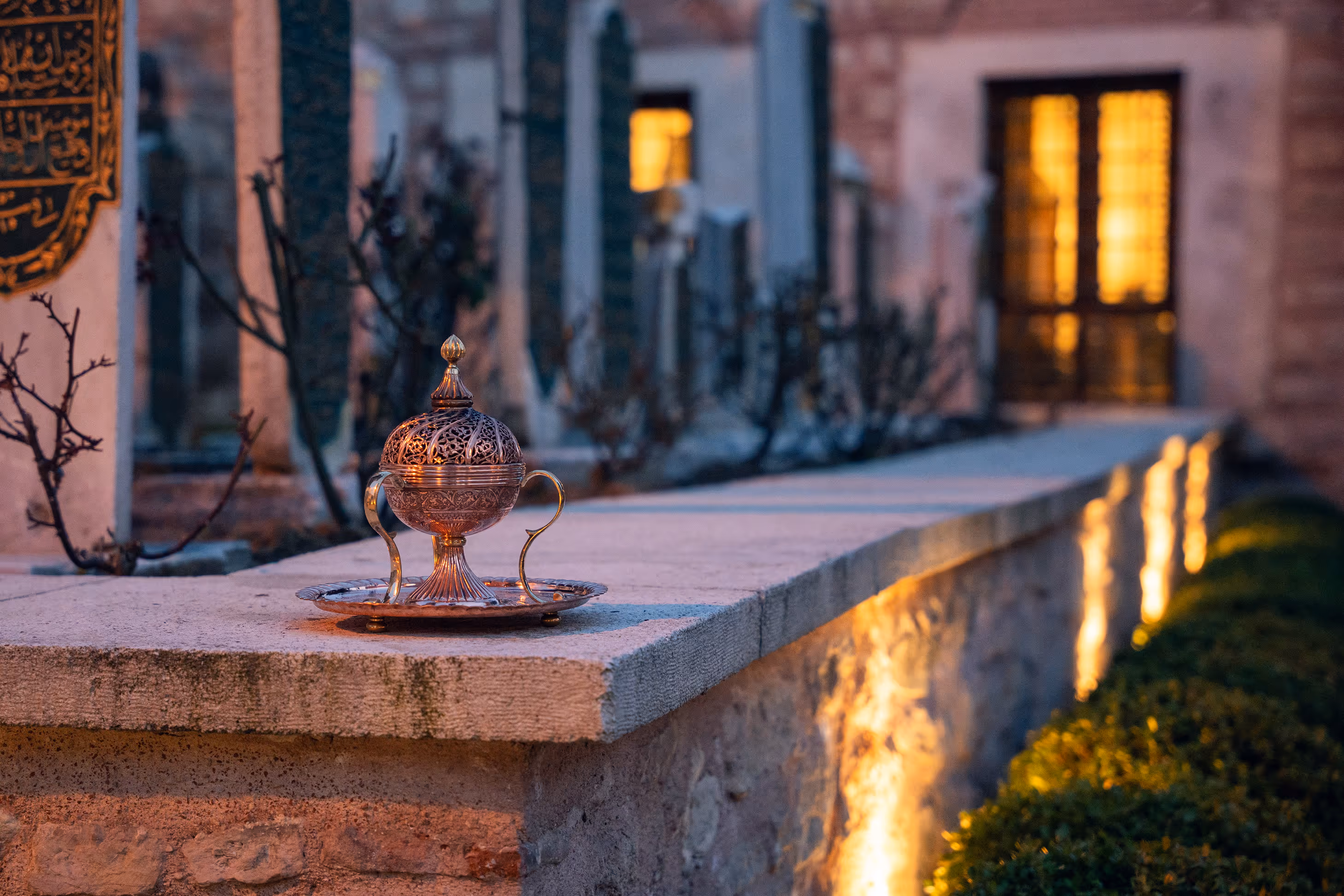 Ornate metal incense burner on a stone ledge with decorative plaques and warm lighting in the background.