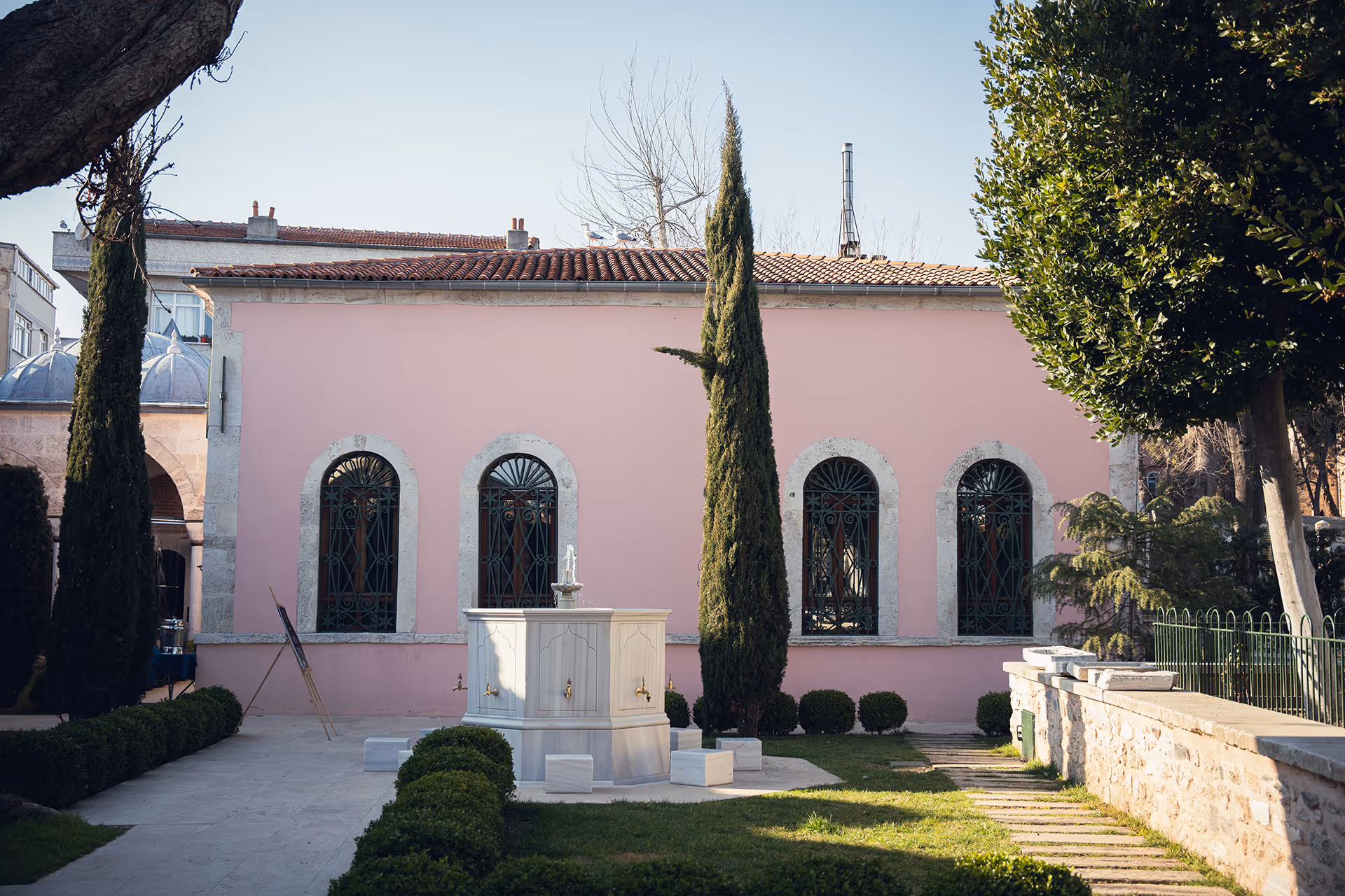 Pink building with four arched windows and decorative ironwork, a small fountain in front surrounded by greenery and tall trees.