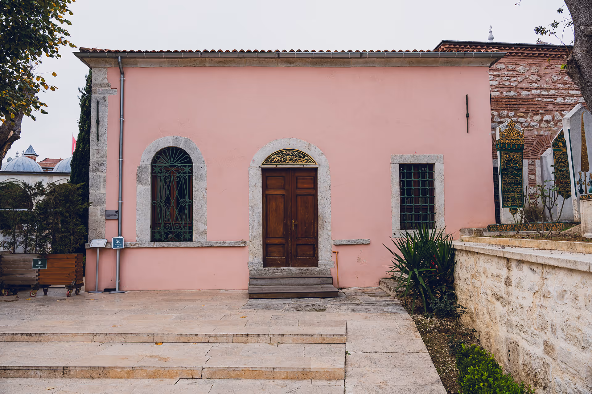 Pink building with wooden double doors, arched windows with green metal grilles, and a small garden beside stone steps.