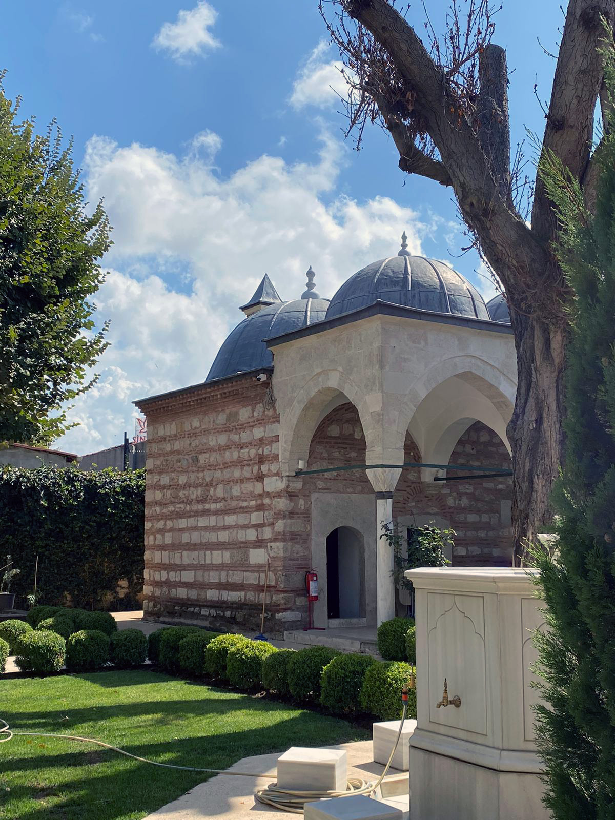 Stone building with domed roofs and arched entrance surrounded by greenery under a blue sky.