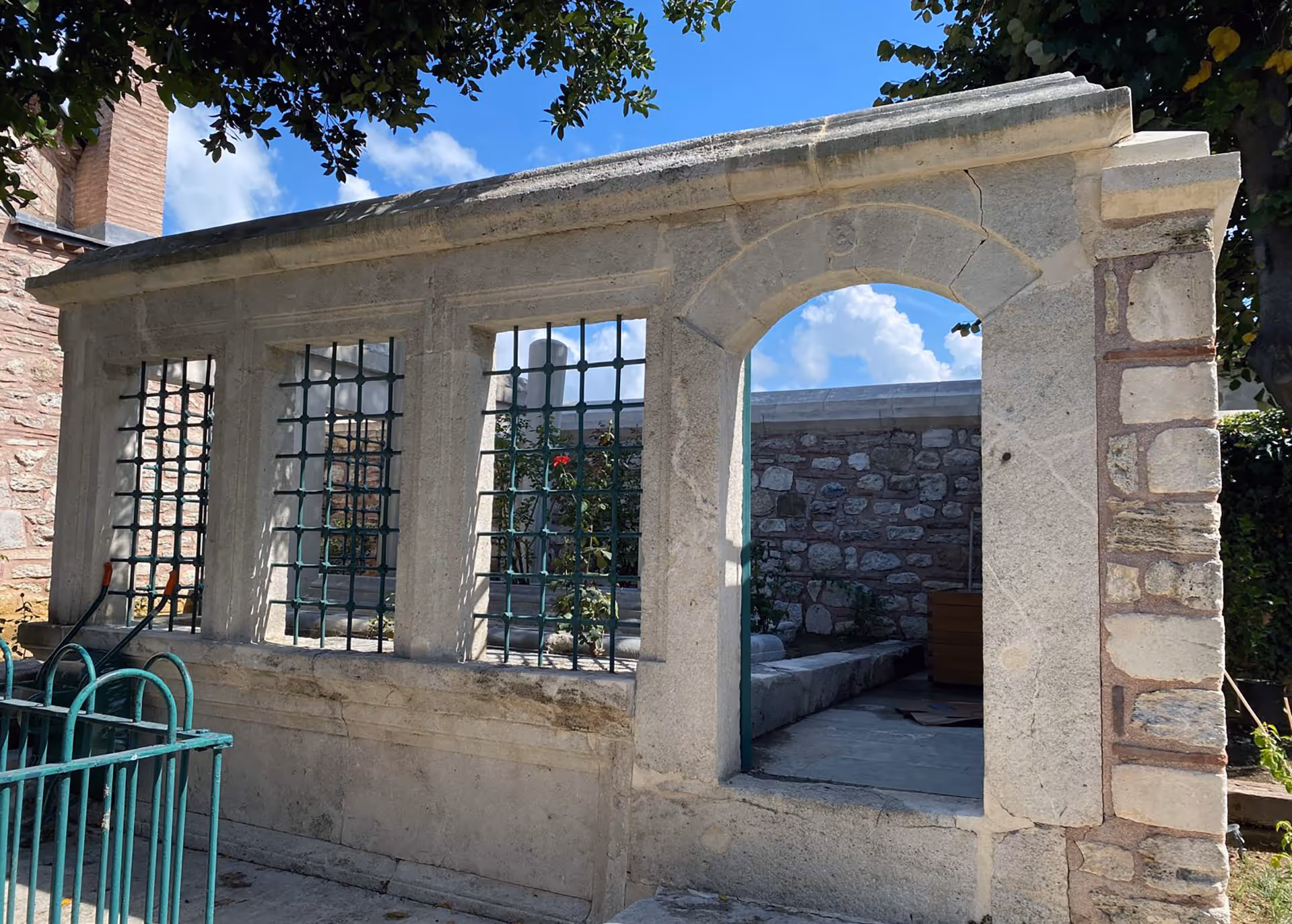 Stone structure with barred windows and an open archway, showing a small courtyard with stone walls and a flower.