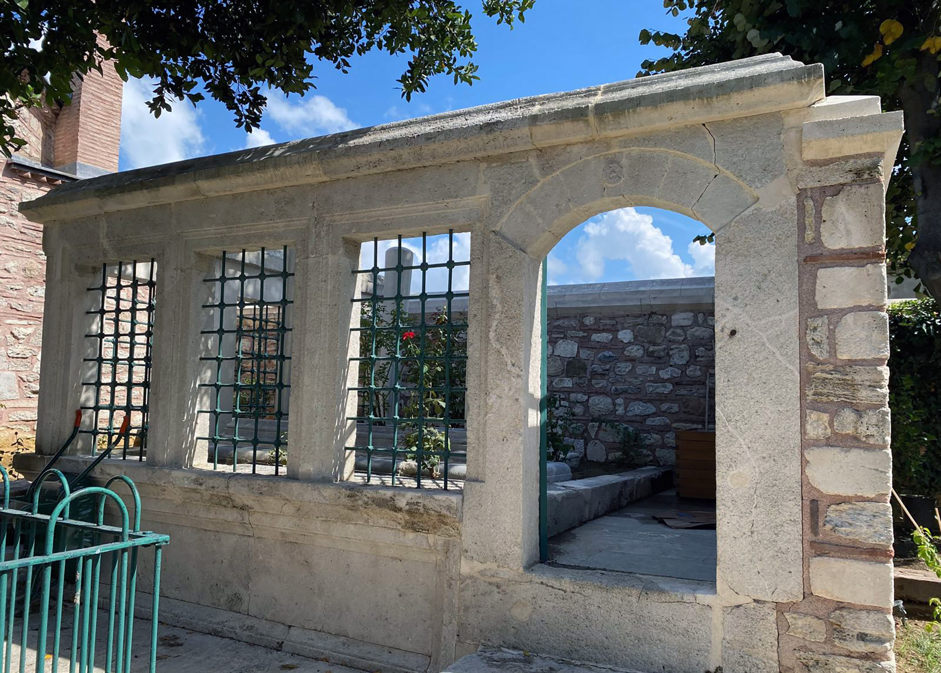 Stone structure with barred windows and an open archway, showing a small courtyard with stone walls and a flower.