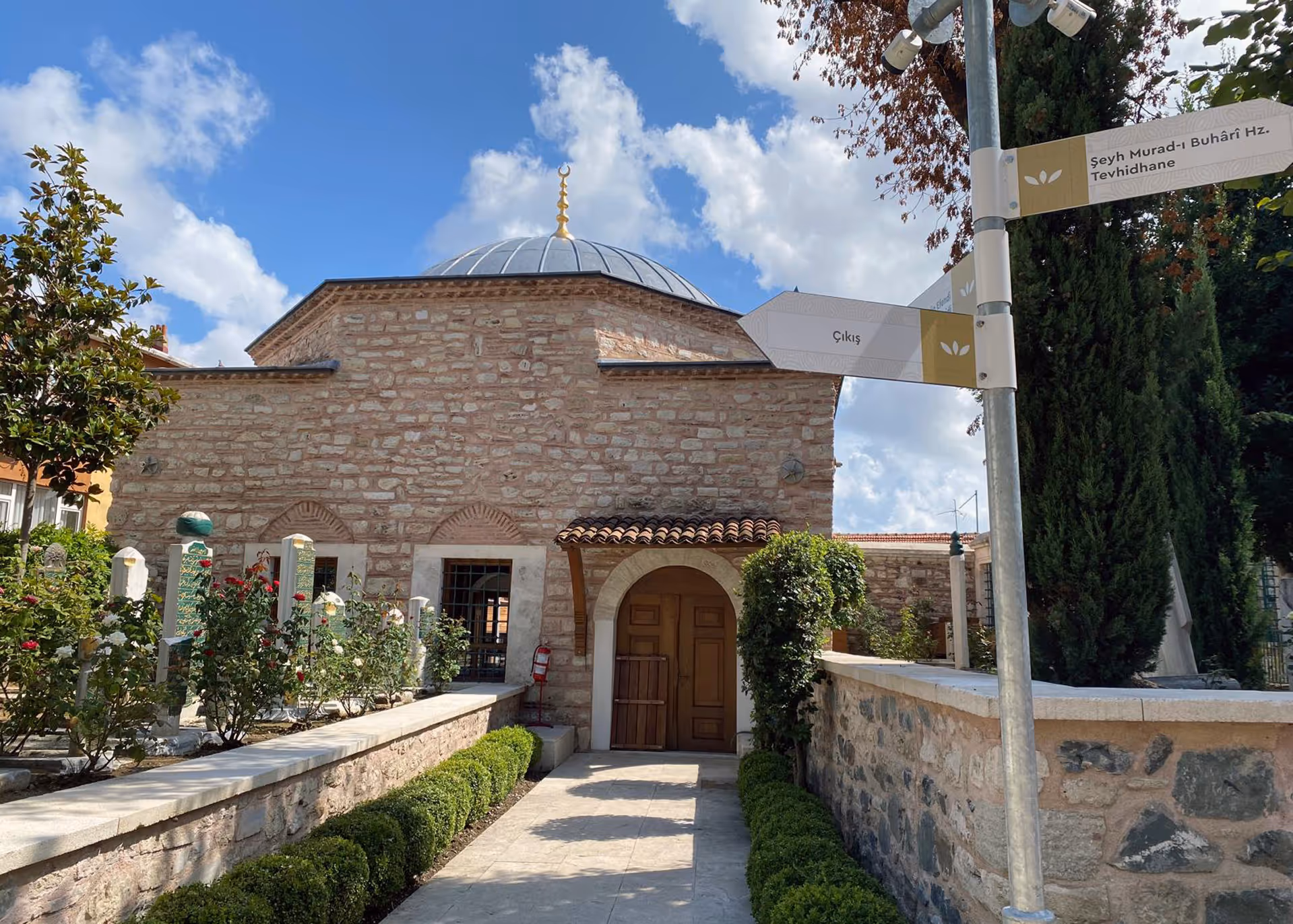 Stone building with a domed roof topped with a crescent finial, surrounded by a garden with rose bushes and signposts in Turkish.