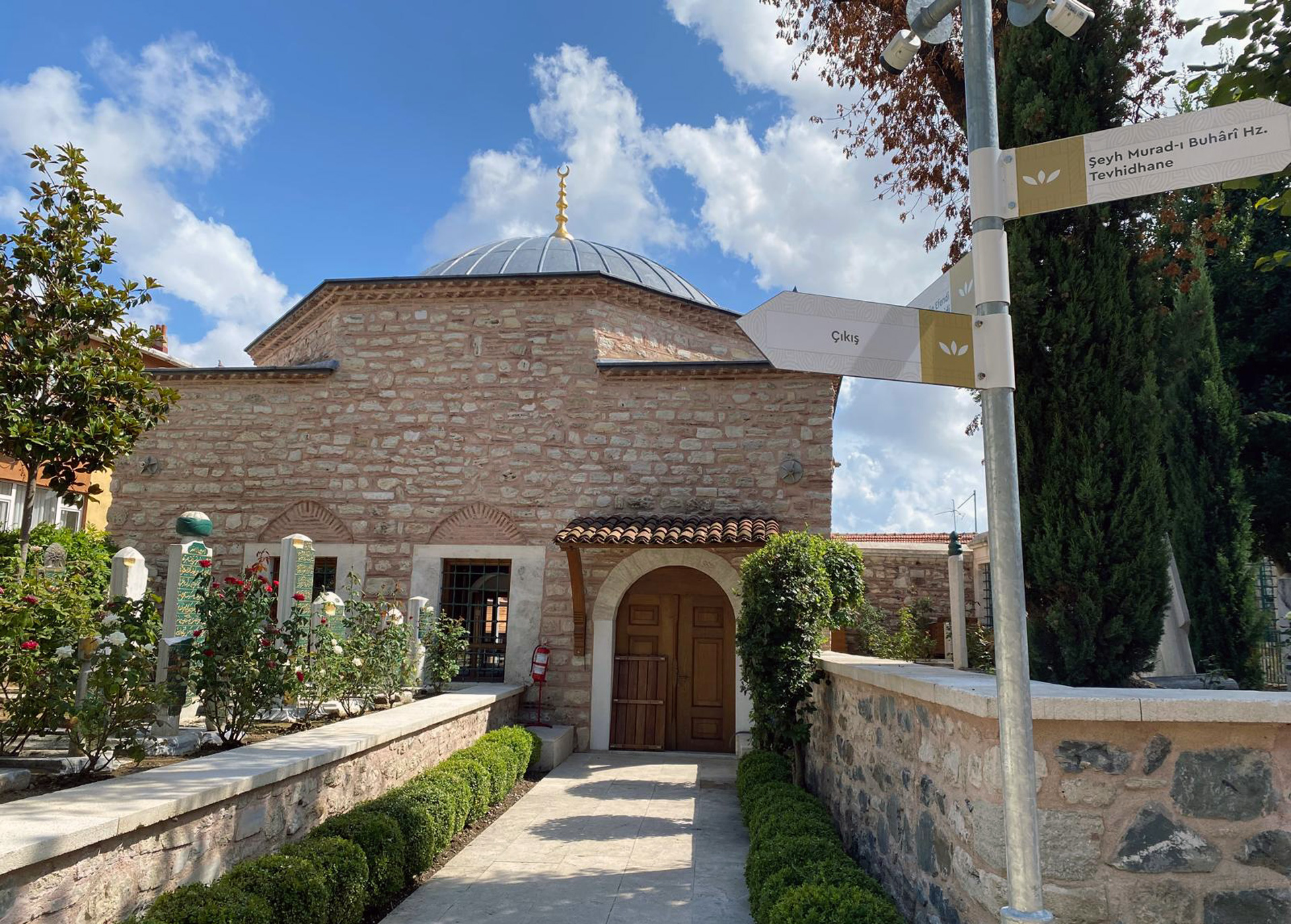 Stone building with a domed roof topped with a crescent finial, surrounded by a garden with rose bushes and signposts in Turkish.