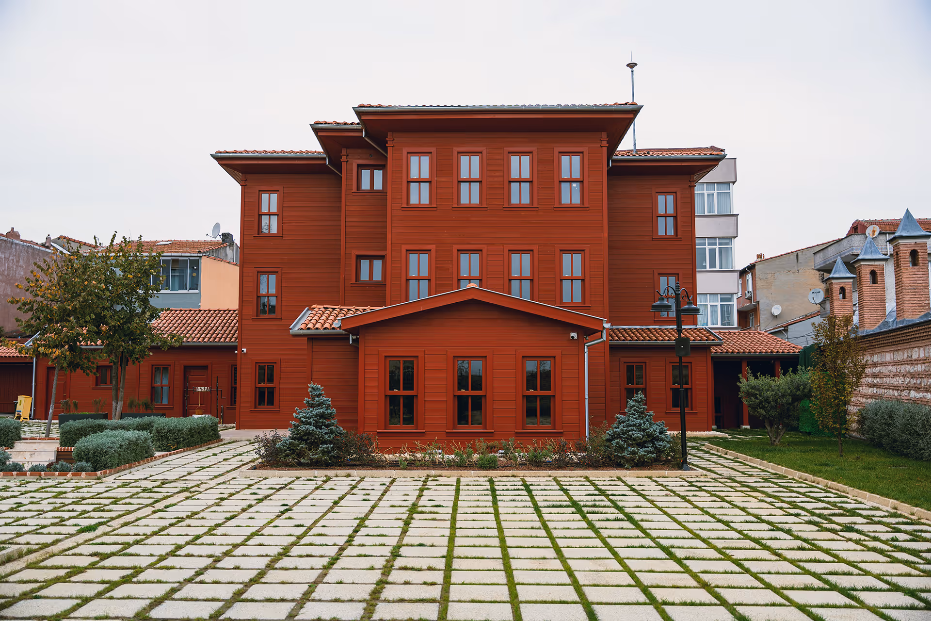 Large red wooden building with multiple windows, a tiled roof, and landscaped paved courtyard with shrubs and small trees.