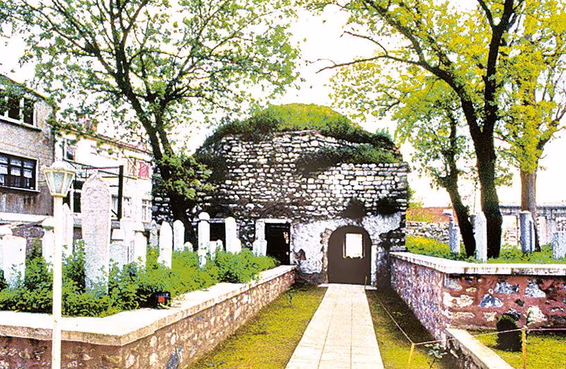 Stone structure with arched doorways surrounded by cemetery with white tombstones and trees.