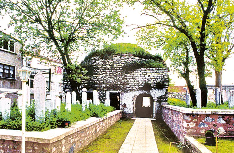 Stone structure with arched doorways surrounded by cemetery with white tombstones and trees.