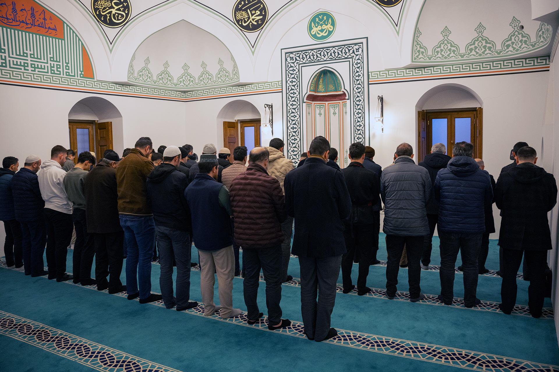Men standing in rows praying inside a mosque with ornate Arabic calligraphy and geometric patterns on the walls.