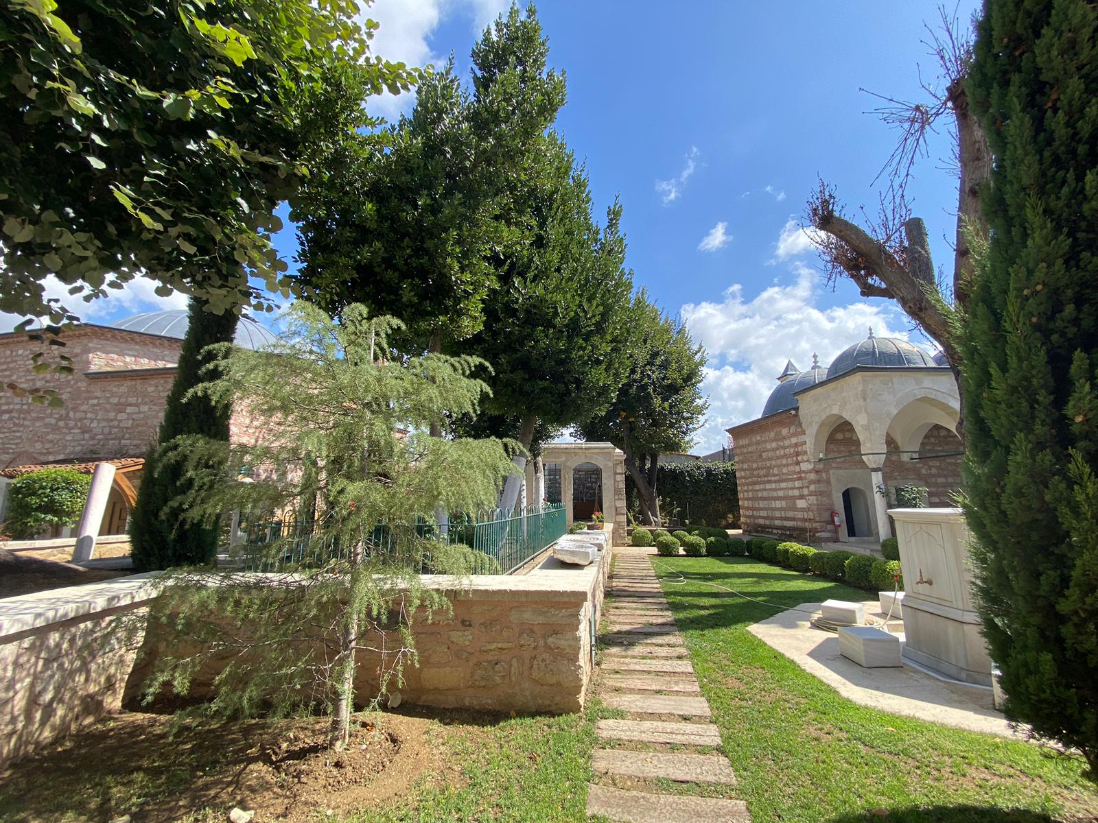 Sunlit garden with trimmed bushes, trees, stone pathways, and historic stone buildings with domed roofs under a blue sky with clouds.