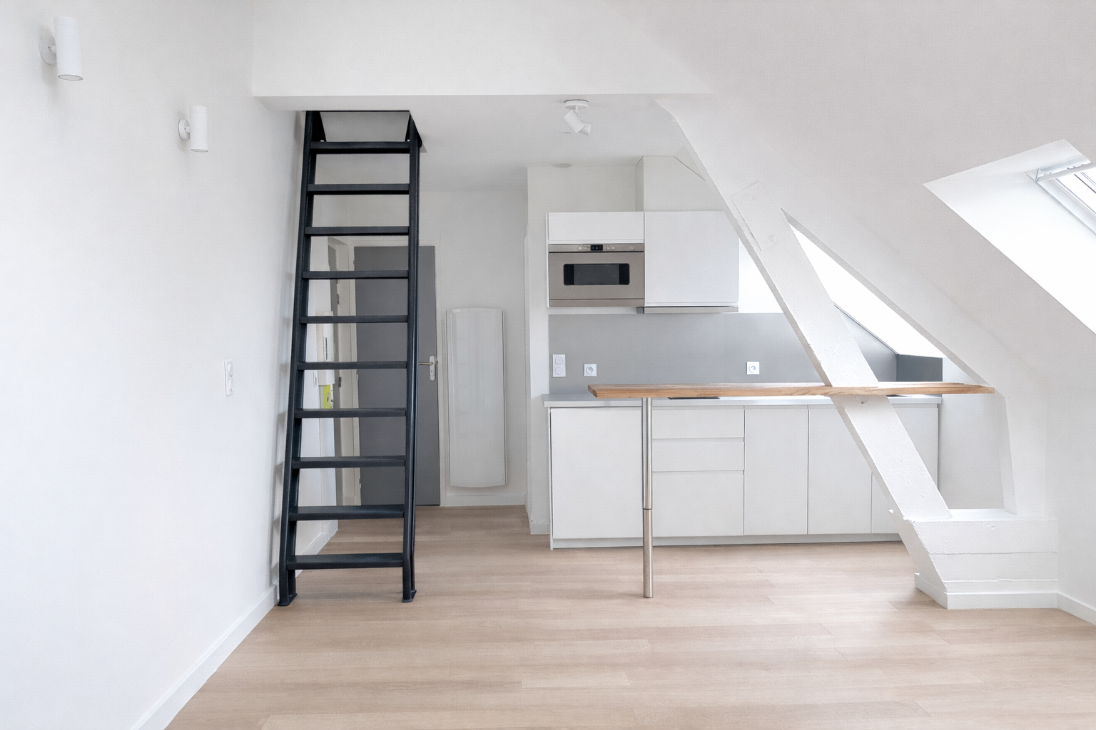 Cuisine moderne minimale avec plancher en bois clair, escalier noir étroit menant à une mezzanine et large poutre blanche inclinée sous une fenêtre de toit.