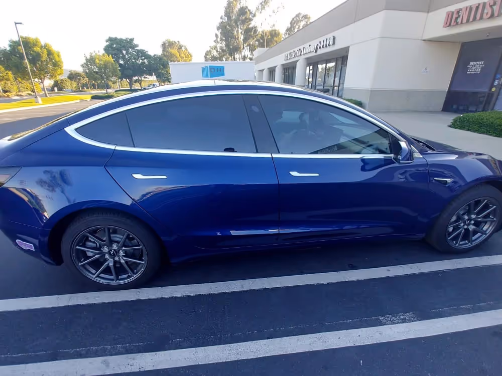 Side profile of a deep blue Tesla Model 3 electric car with tinted windows parked in a sunny outdoor parking lot.