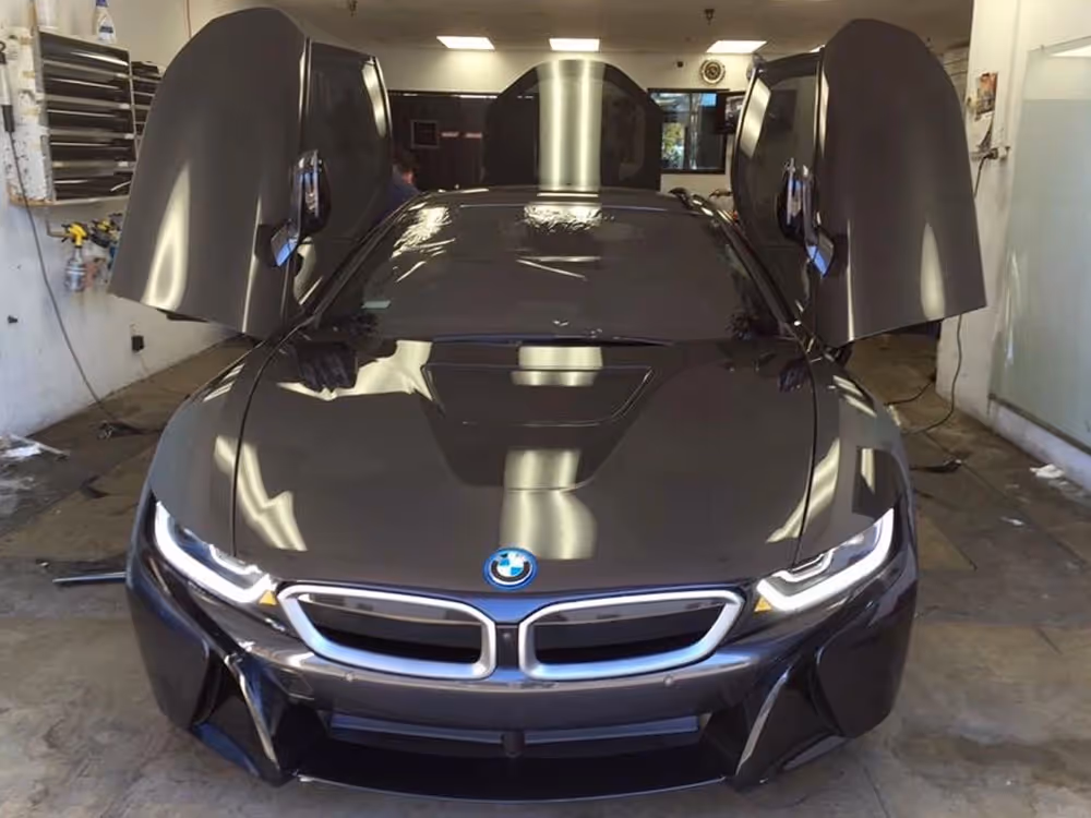 Black BMW i8 hybrid sports car with butterfly doors open, illuminated LED headlights, and iconic kidney grille in a garage.