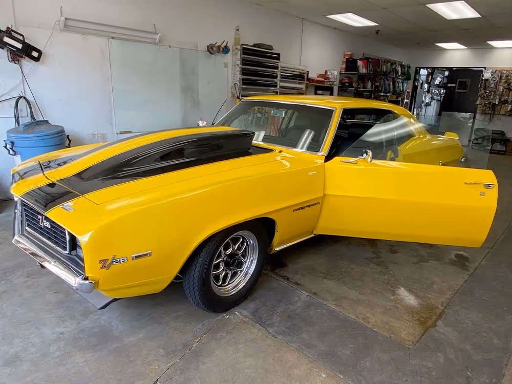Yellow 1969 Chevrolet Camaro muscle car with black racing stripes and open door in a professional auto shop.