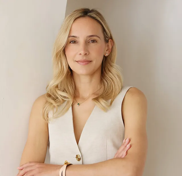 Emily with shoulder-length hair wearing a sleeveless beige top, standing with arms crossed against a light background.