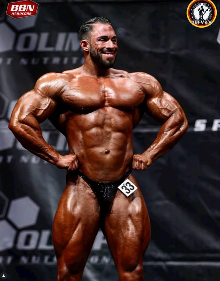 Lachender Mann mit Tattoos auf Oberkörper und Armen erhält eine Schultermassage von einer Person in schwarzem T-Shirt.