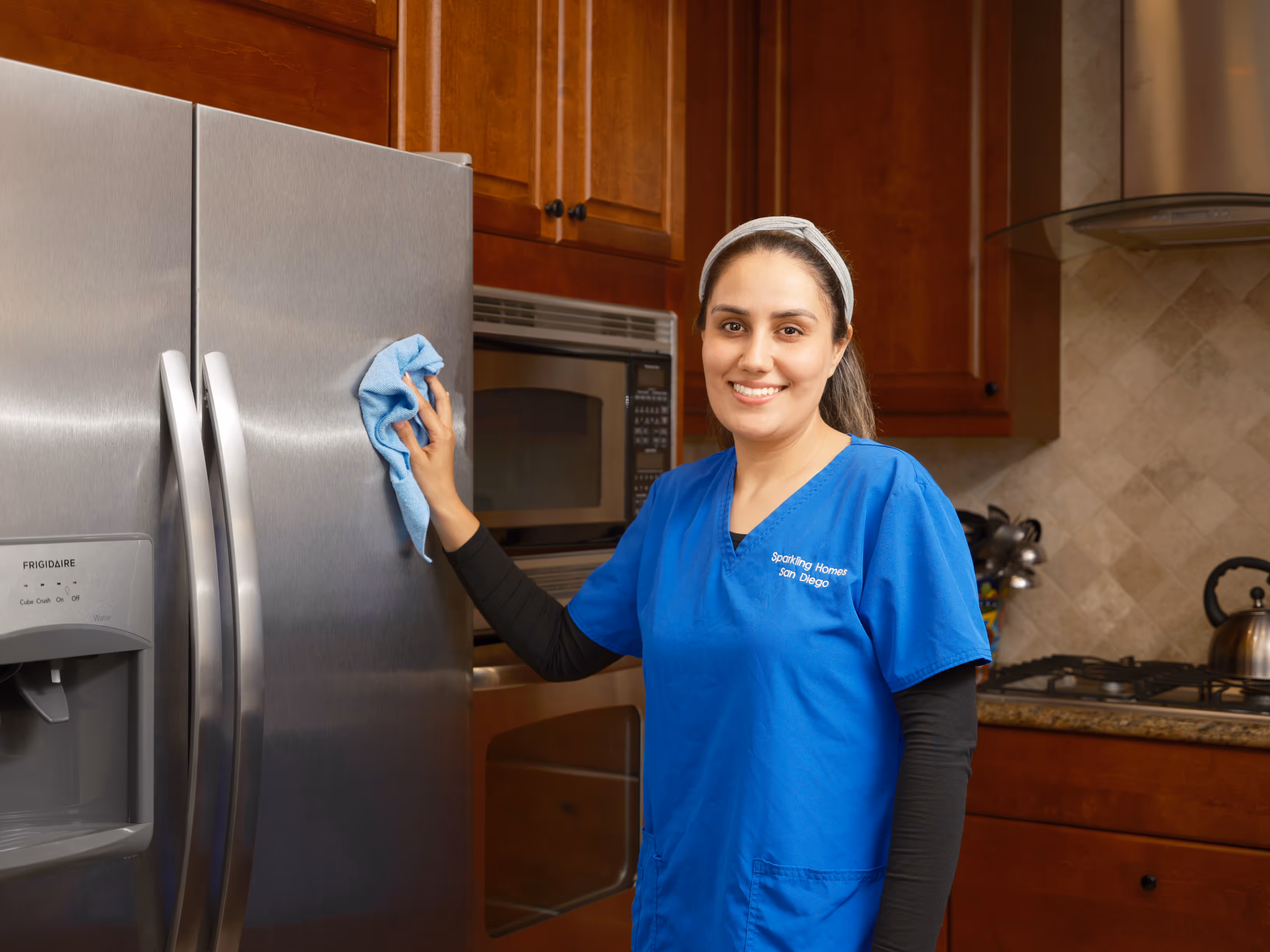 Woman in blue cleaning uniform wiping a stainless steel refrigerator door with a cloth in a kitchen.