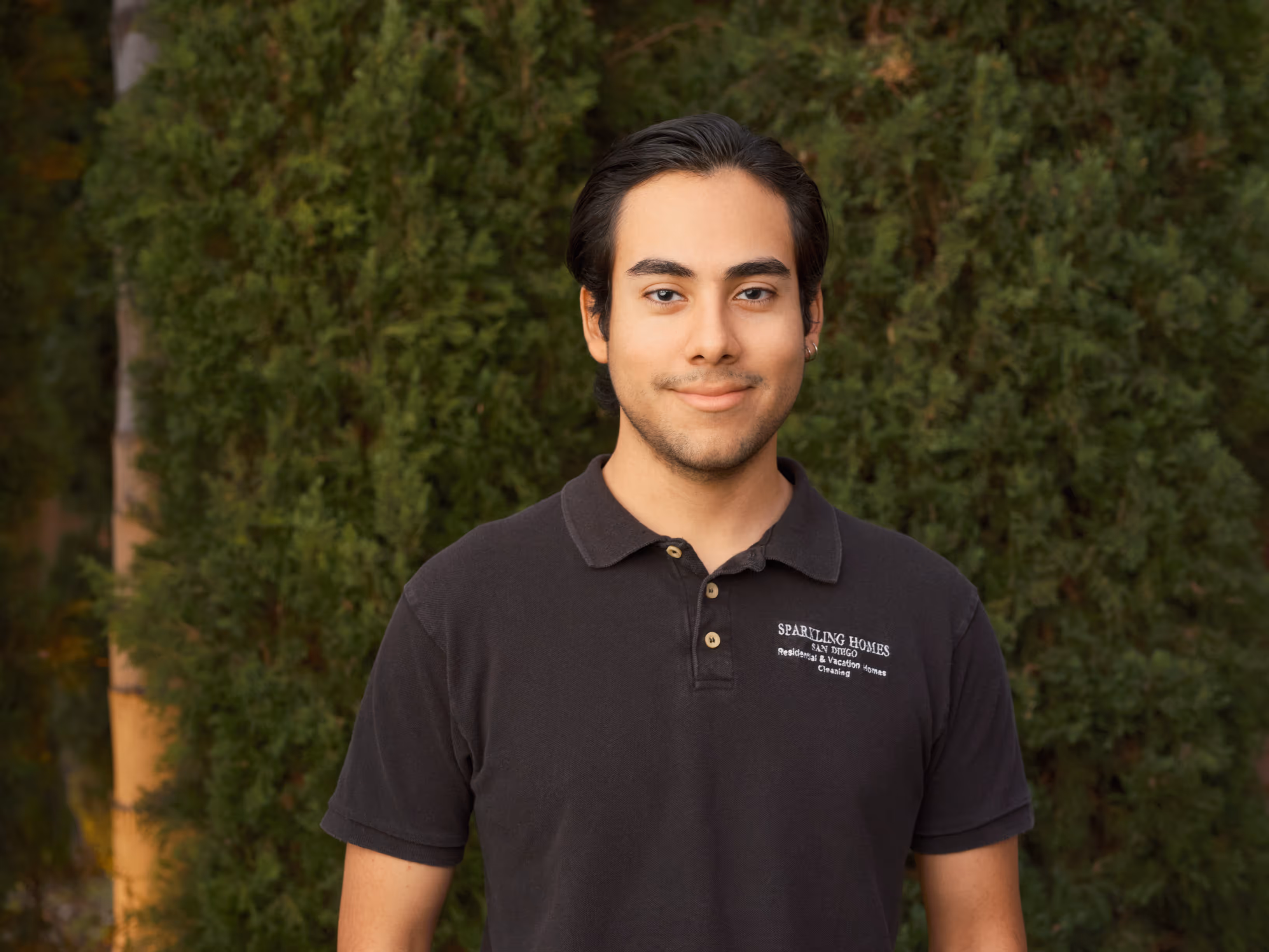 Young man with dark hair wearing a black polo shirt with Sparkling Homes logo, standing in front of green foliage.