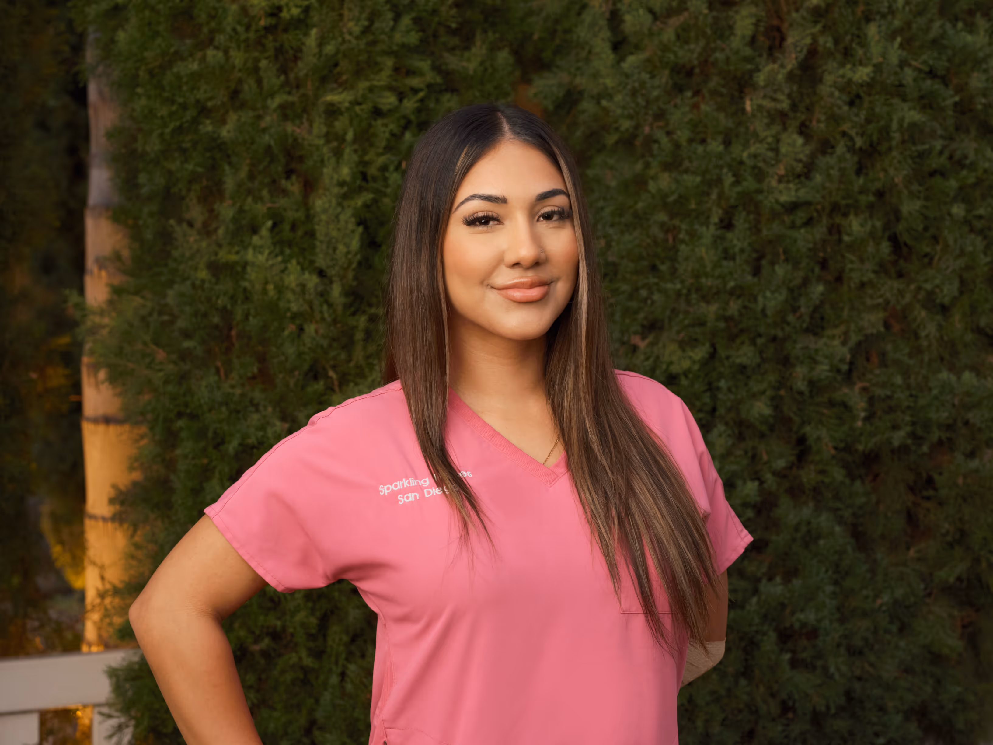 Woman with long brown hair wearing a pink medical scrub top, standing outdoors in front of greenery and smiling.