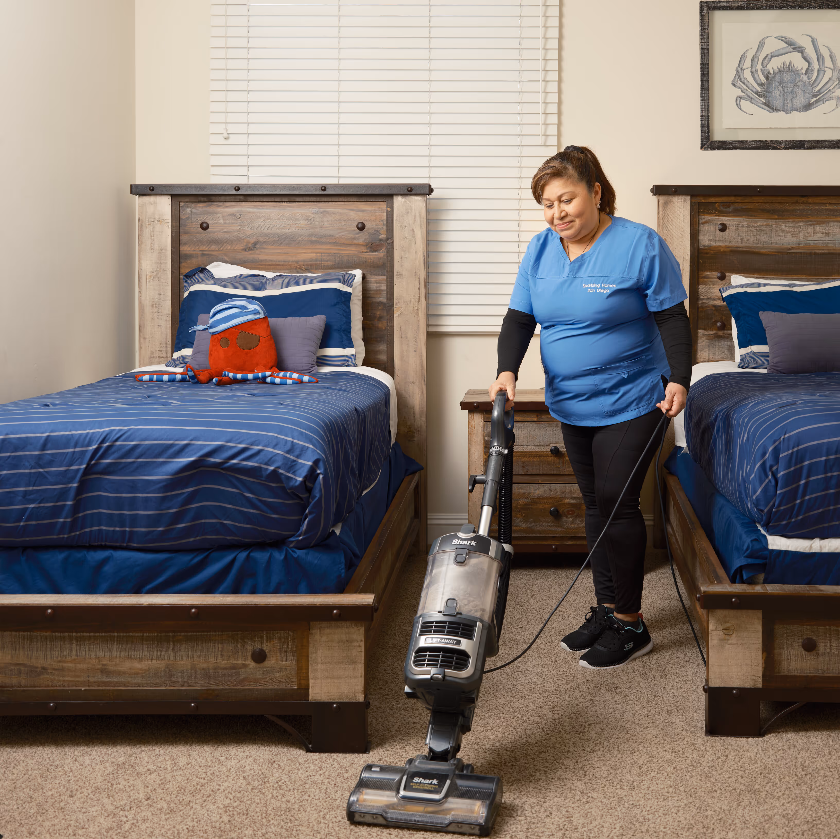 Woman in blue scrubs vacuuming carpet between two beds with blue-striped bedding in a bedroom.