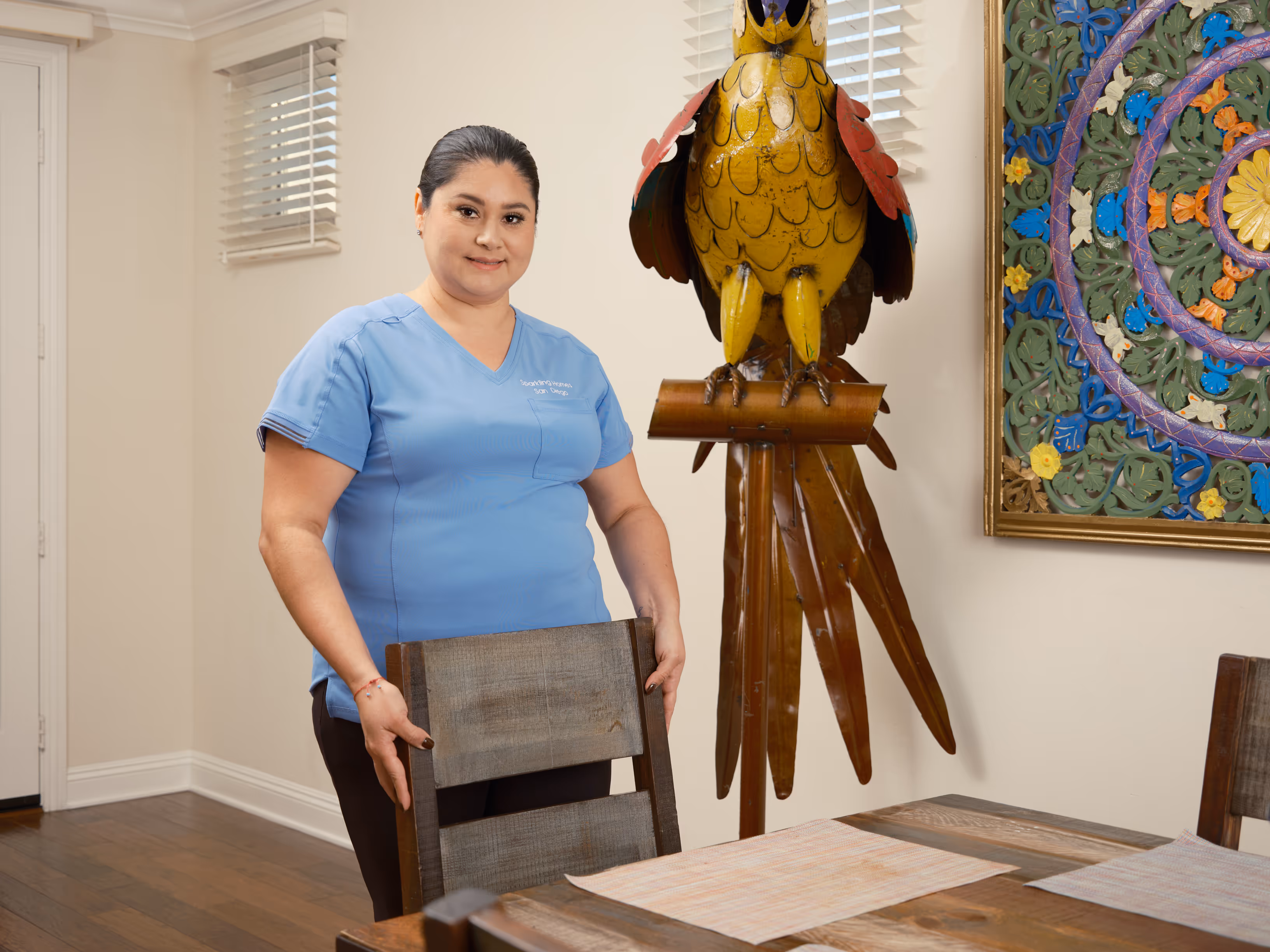 Woman in blue scrubs standing behind a wooden chair in a dining room with a large yellow bird sculpture and colorful wall art.