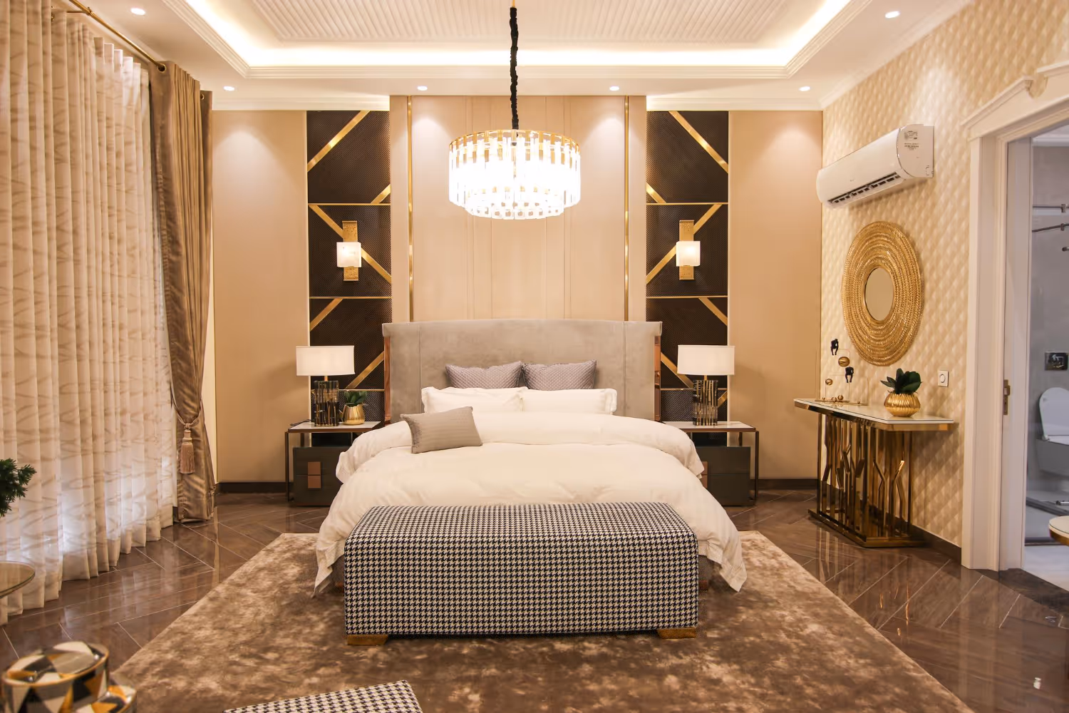 Luxurious bedroom with a large bed, beige headboard, patterned bench, golden accents, and a chandelier.