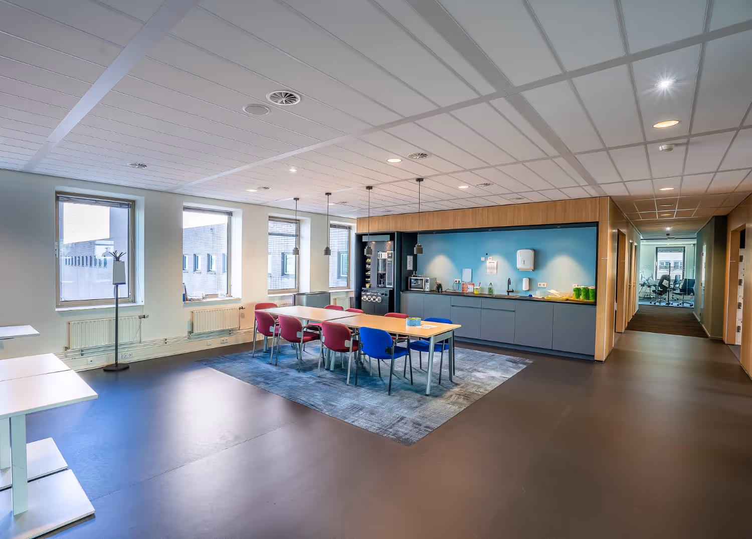 Modern office break room with a long table surrounded by red and blue chairs, kitchen area with cabinets, and large windows.