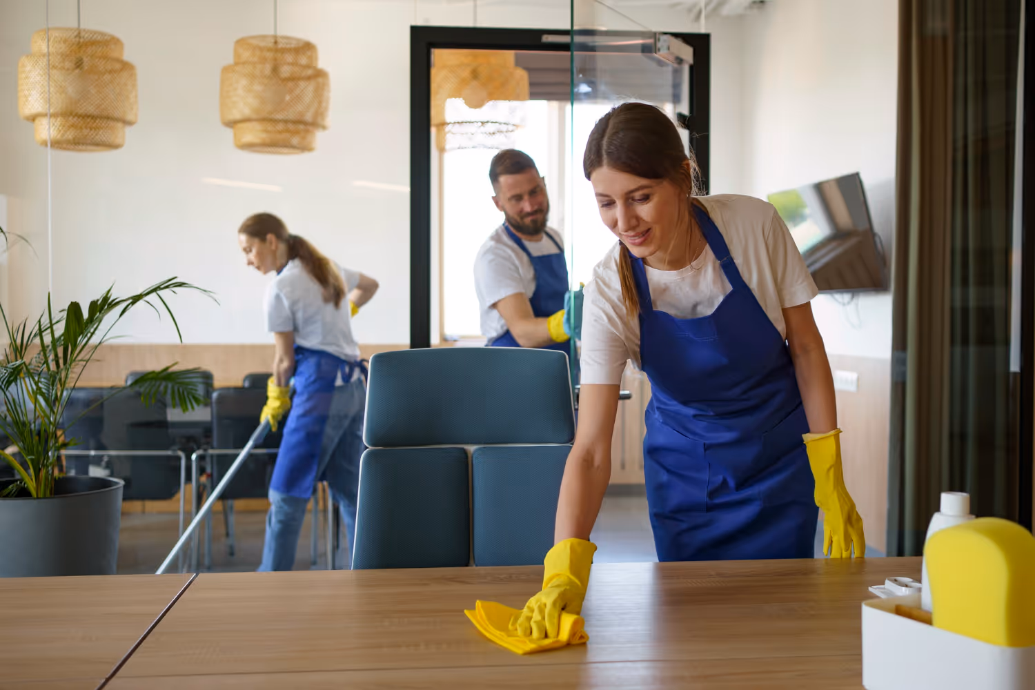 Three professional cleaners in yellow gloves and blue aprons cleaning an office, wiping a table, mopping the floor, and cleaning a glass wall.