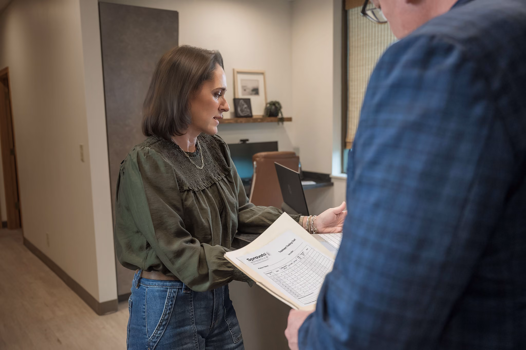 Woman holding a folder and talking to a man in a plaid jacket in an office setting.