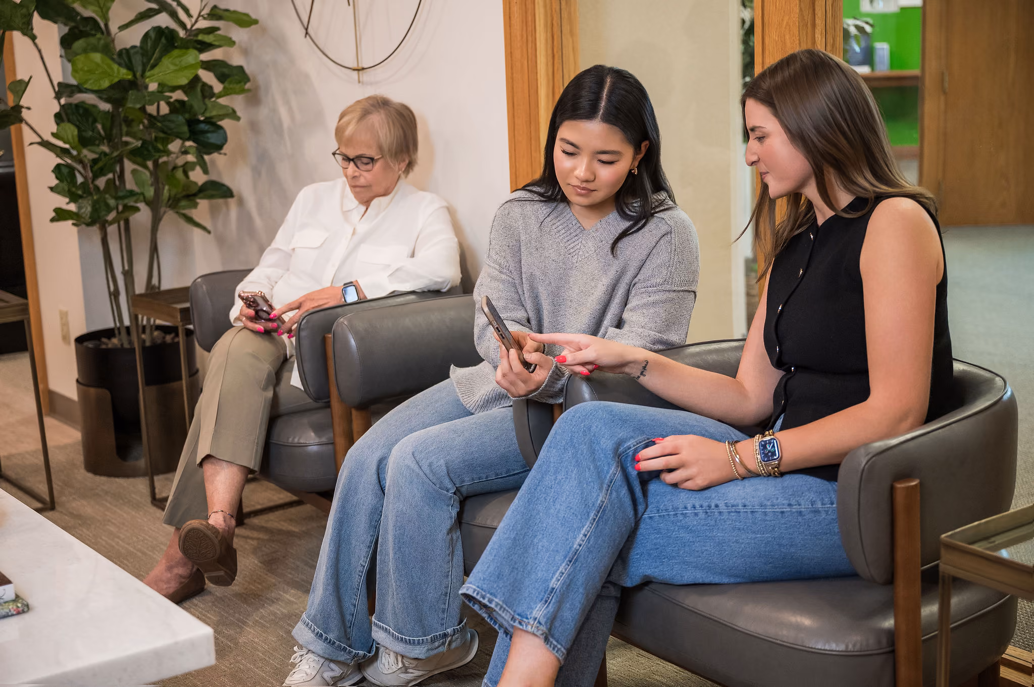 Three women seated indoors: one older woman looks at phone, two younger women share a phone.