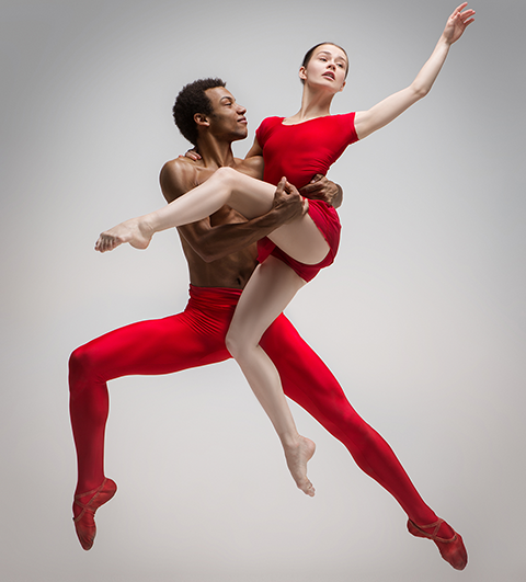 Dancer in a gray sweater and bow tie performing a wide dance stretch against a textured brown wall.