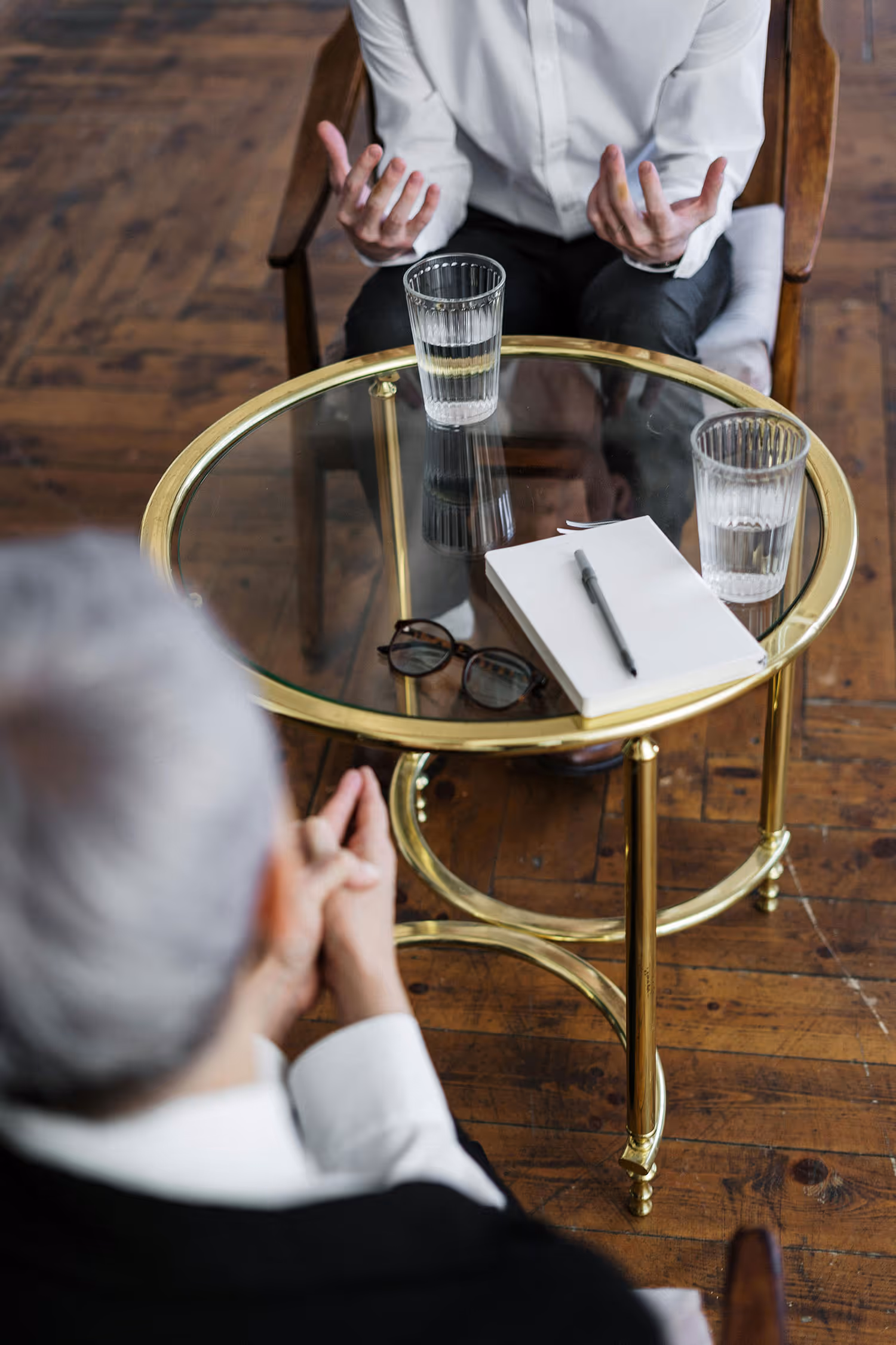 Two people seated across from each other at a glass-top metal table with two glasses of water, a notebook, a pen, and glasses on the table.