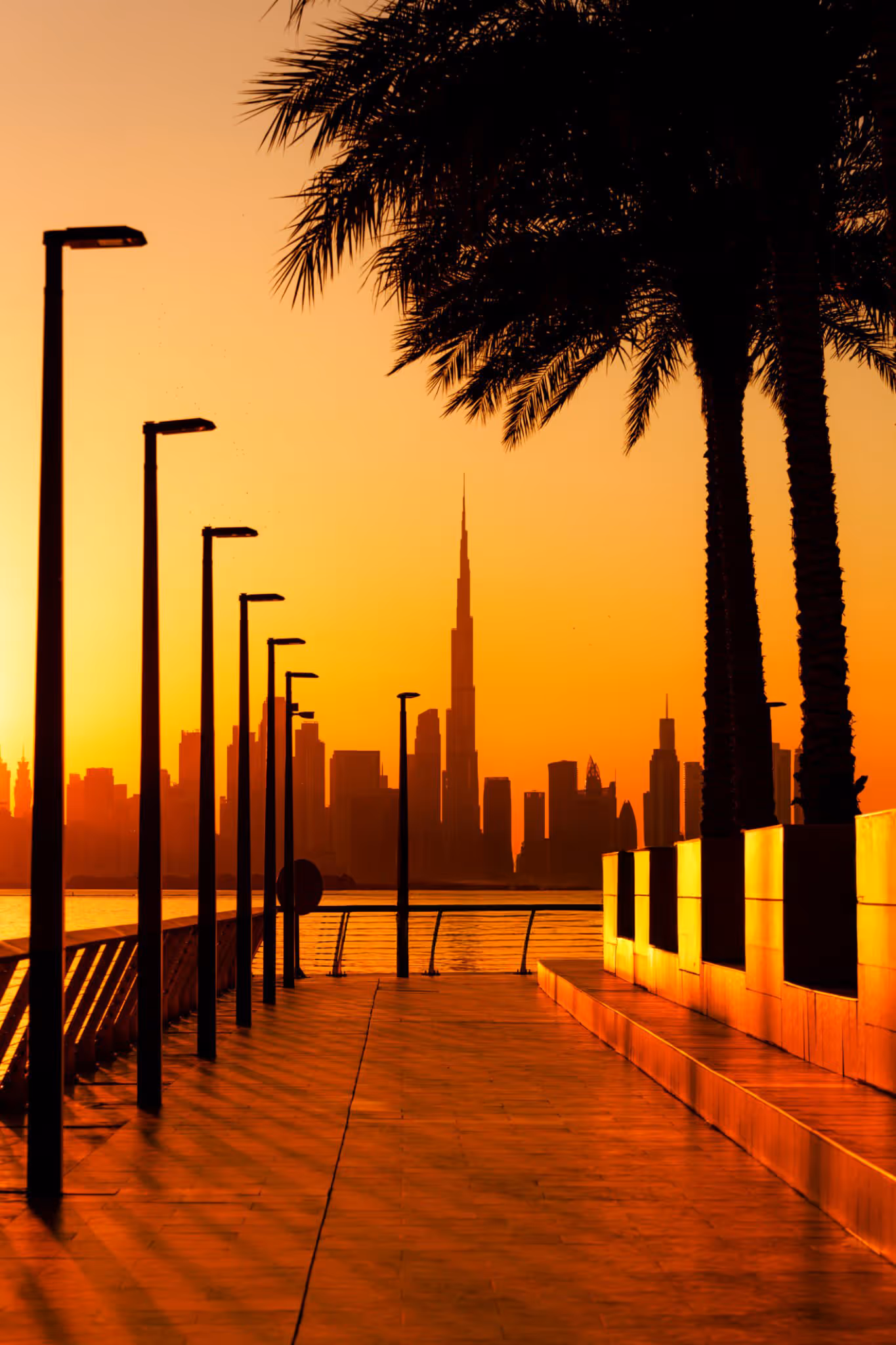 Sunset view of Dubai skyline with Burj Khalifa in the center, silhouetted palm trees, and walkway with street lamps.