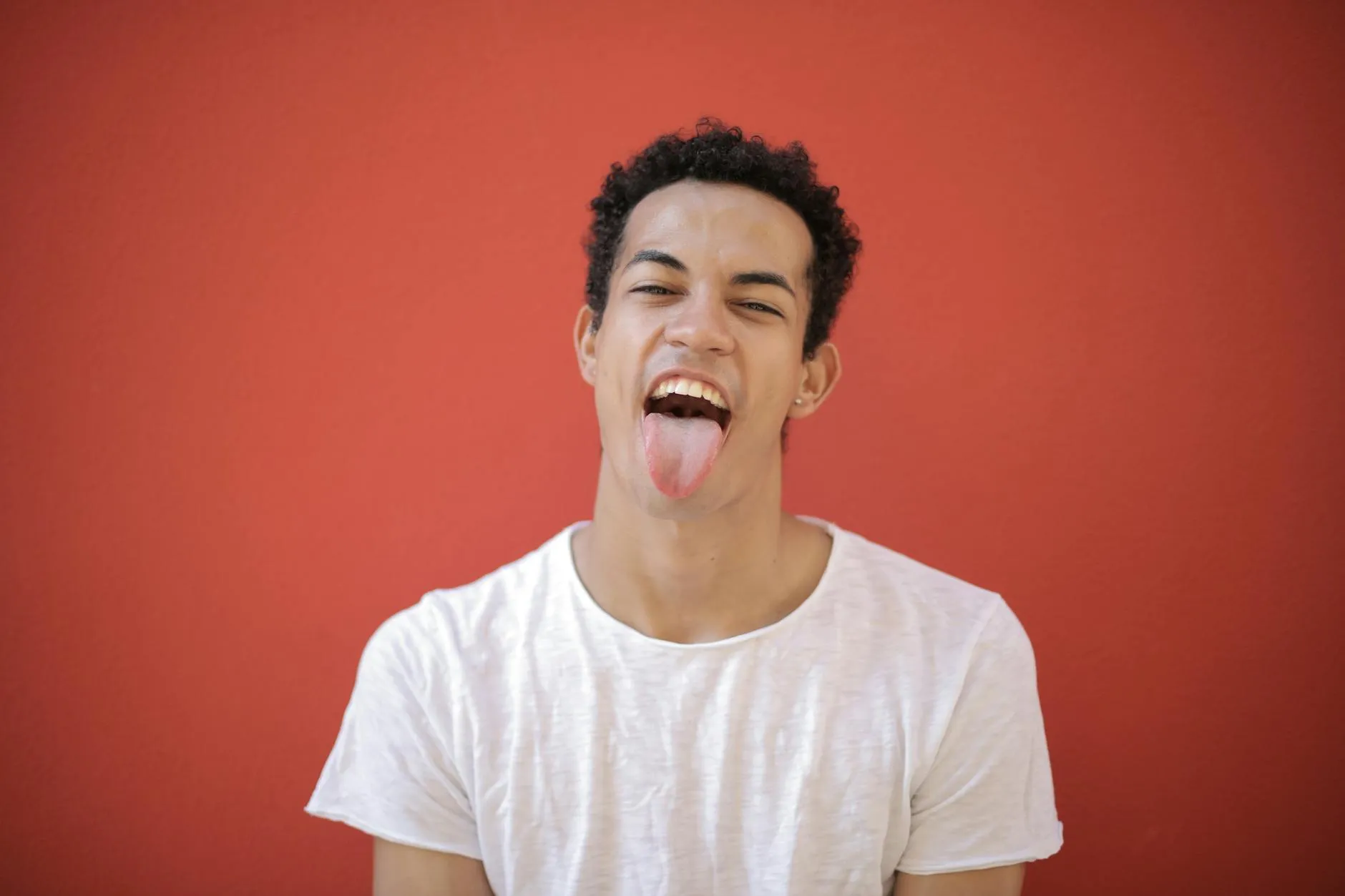 Young man in a white t-shirt sticking out his tongue and smiling playfully against a red background.