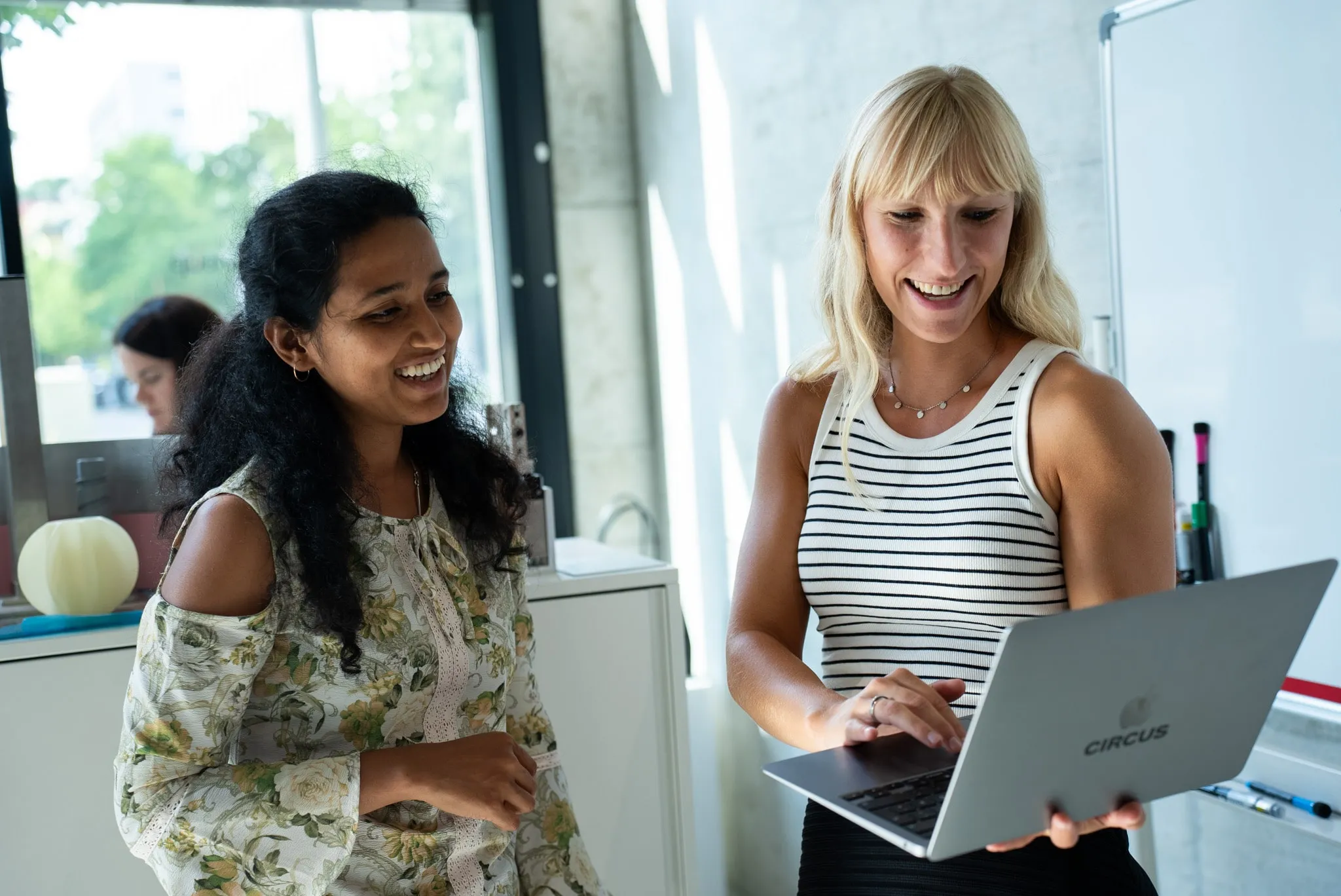 Two women smiling and collaborating while looking at a Circus-branded laptop in a bright office space.