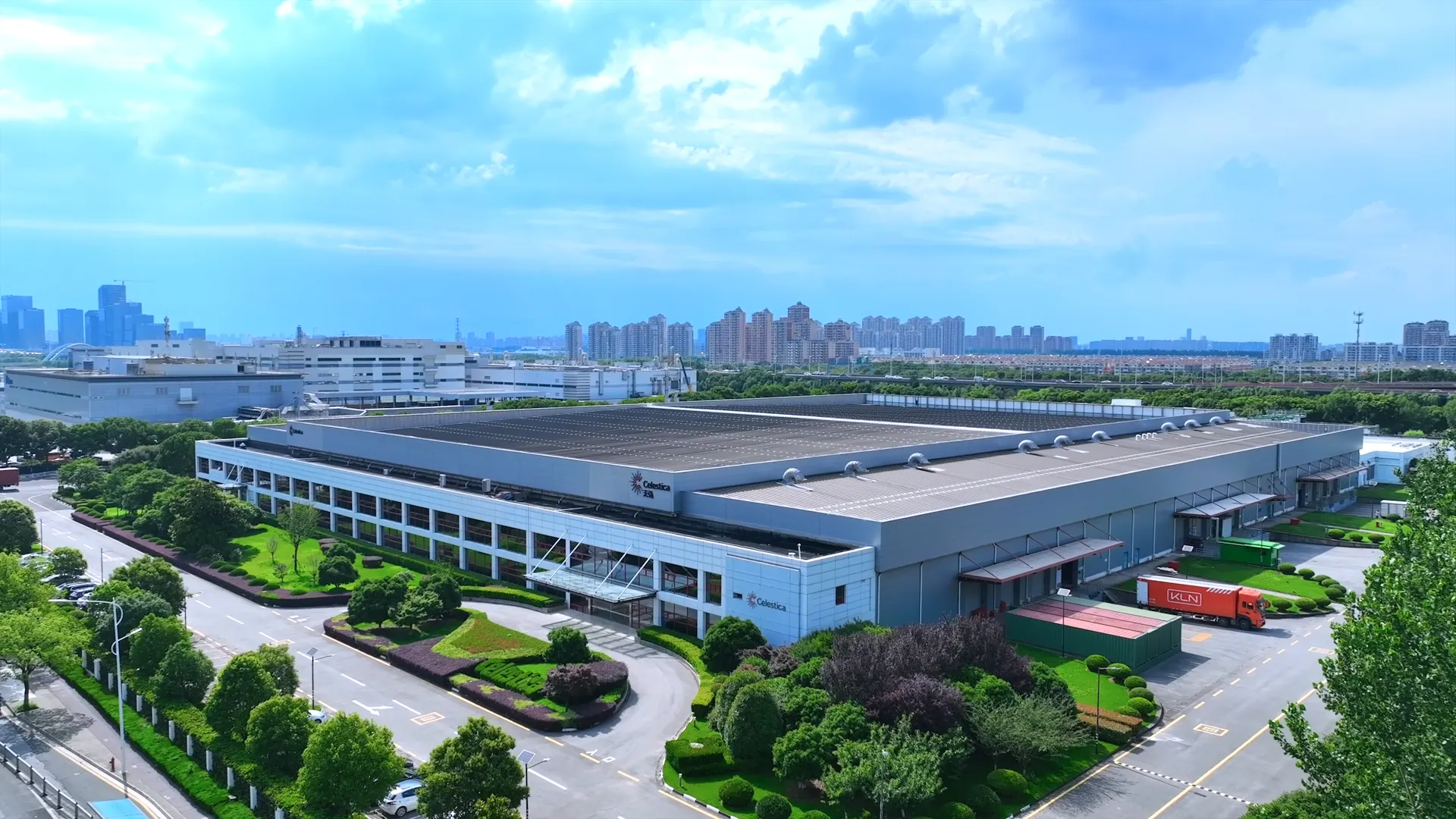 Aerial view of a large industrial building with Celestica signage, surrounded by greenery and roads, with a city skyline in the background under a partly cloudy sky.
